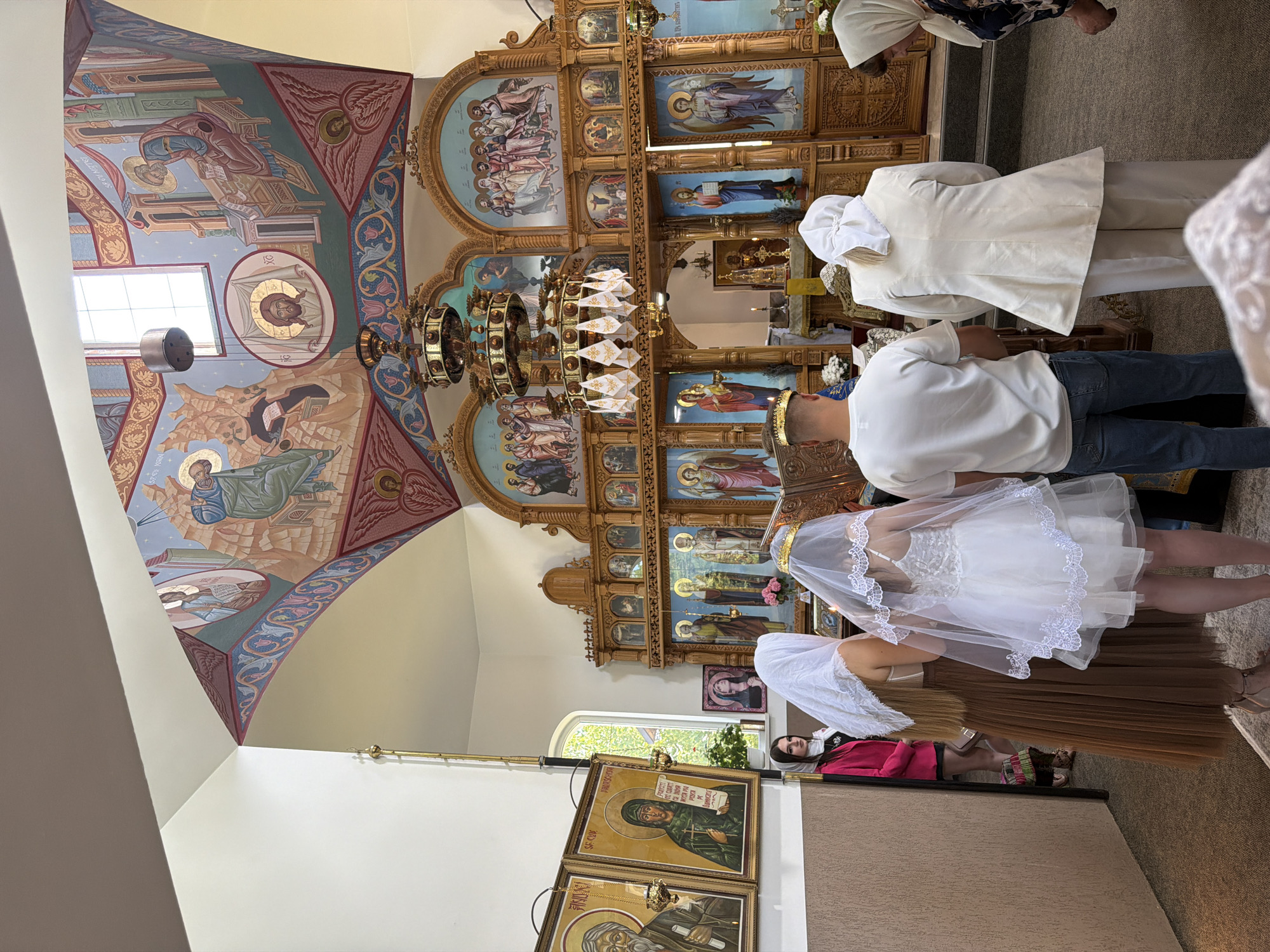 The image depicts the interior of an Orthodox church in Strășeni, Moldova. In the foreground, a group of people stand facing an ornate wooden iconostasis, a wall of icons and religious imagery. A young couple, presumably the bride and groom, are central to the scene, both wearing crowns, indicating a wedding ceremony. The bride wears a white veil and a short white dress, while the groom is in a white t-shirt and jeans. Another woman in a white veil and a brown skirt is positioned to the left of the couple. Further to the left, a woman in a bright pink jacket and a dark skirt is visible near a doorway. The iconostasis itself is richly decorated with multiple rows of religious icons and carved wooden details. Above the iconostasis, the ceiling is adorned with religious murals and a large, decorative chandelier hangs in the center of the room. The overall atmosphere appears to be one of solemnity and religious observance, consistent with a church ceremony.
