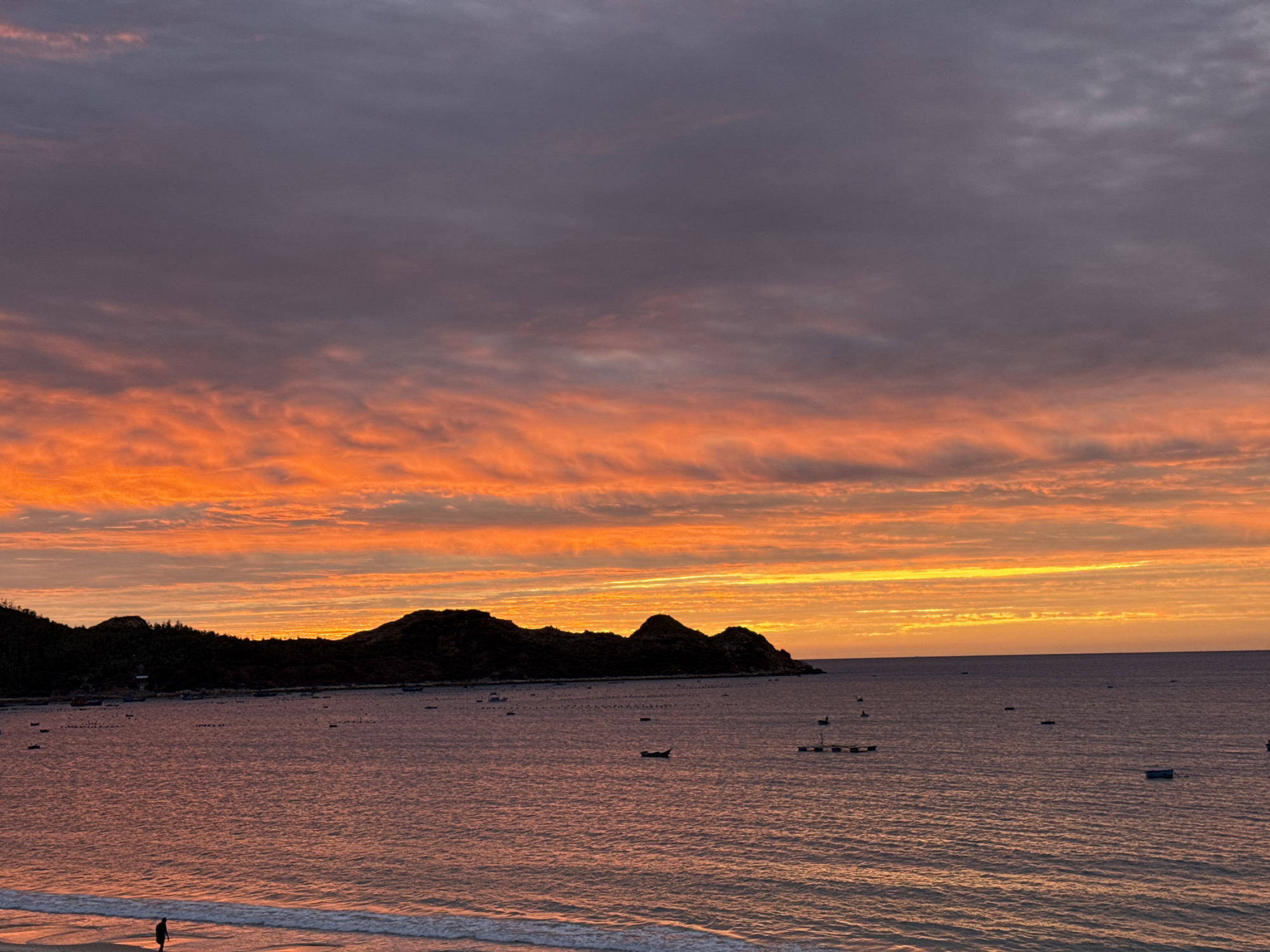 A tranquil ocean scene unfolds at dusk, with a dramatic sky ablaze in fiery oranges and yellows near the horizon, transitioning to deep purples and grays overhead. The sun, not directly visible, casts a warm, diffused glow that reflects on the rippling water. In the distance, a silhouetted island or peninsula with undulating hills is visible along the coastline, dotted with a dark line of trees. Several small boats are scattered across the water, some appearing to be moored or anchored. A sandy beach occupies the foreground, with gentle waves lapping at its edge. A lone figure stands on the beach, facing the ocean, their silhouette dark against the water's reflection. The overall atmosphere is serene and contemplative, capturing the quiet beauty of twilight by the sea.