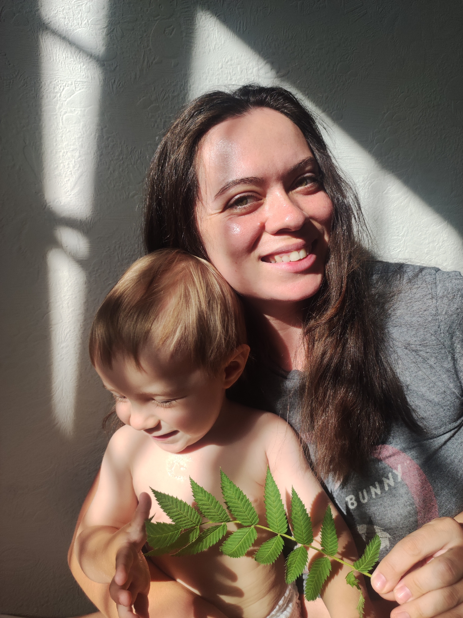 A woman and a young child are close together, with the woman smiling at the camera and the child looking down at a leafy green sprig held in the child's hand. The woman is fair-skinned, with long, dark brown hair, and is wearing a gray t-shirt with the word "BUNNY" visible on the shirt. The child has light skin and blondish hair. The child is holding a green leaf with multiple leaflets along a central stem. Sunlight casts harsh shadows on the wall behind them. The exact location of this image cannot be ascertained from the image and the text prompt. There is insufficient information to confidently place the photo within Chaikovsky, Russia.