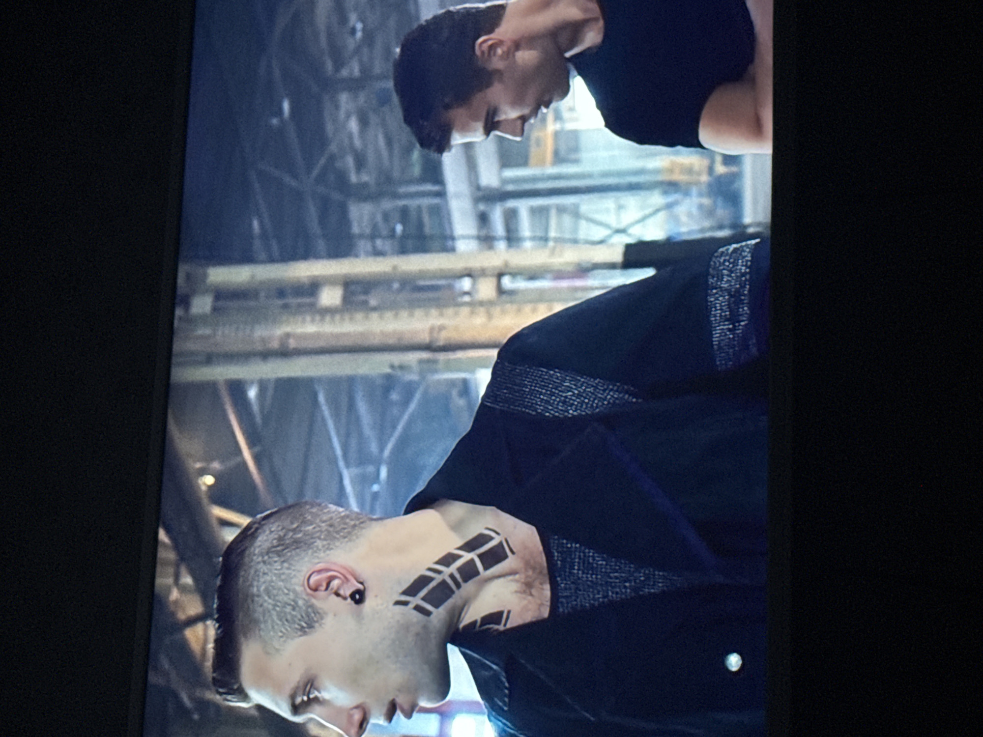 The image shows two men indoors, possibly in an industrial or warehouse-type setting, given the visible structural elements in the background. The man closer to the camera has a short, faded haircut and a black geometric tattoo on his neck. He is wearing a dark blue jacket or robe-like garment. The other man, slightly further away and higher in the frame, has a dark T-shirt. The location is indeterminate due to the limited visual information. Nothing within the image indicates this is within Mytishchi, Russia.