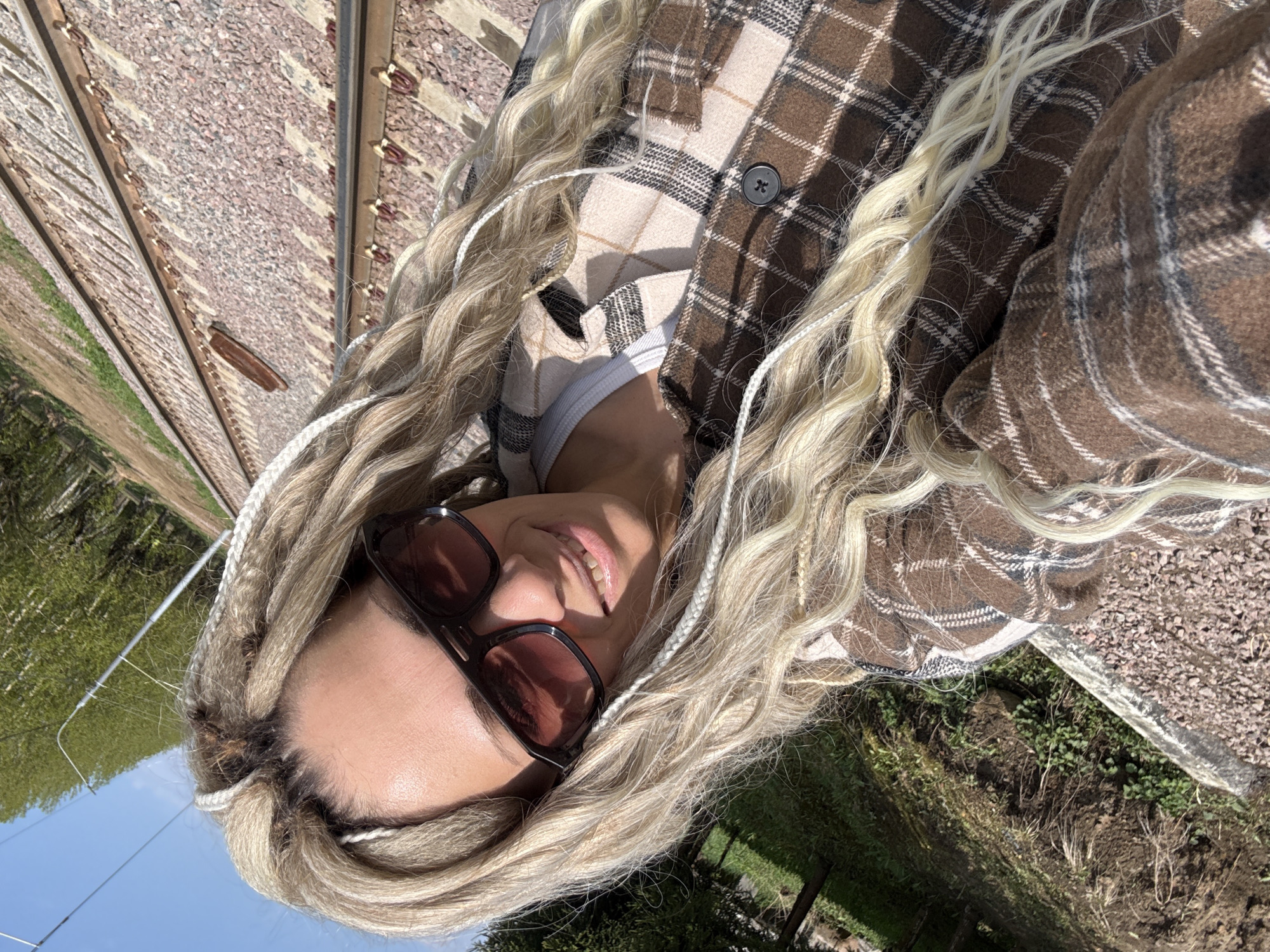 A woman with blonde braided hair and sunglasses is standing near railroad tracks in what appears to be a rural area of Шебунино, Russia. She is wearing a brown and white plaid jacket over a white top. The railroad tracks run parallel to the ground, with gravel and dirt visible around the tracks. Green vegetation and trees are visible in the background. The sky is partially visible with blue patches and sunlight.