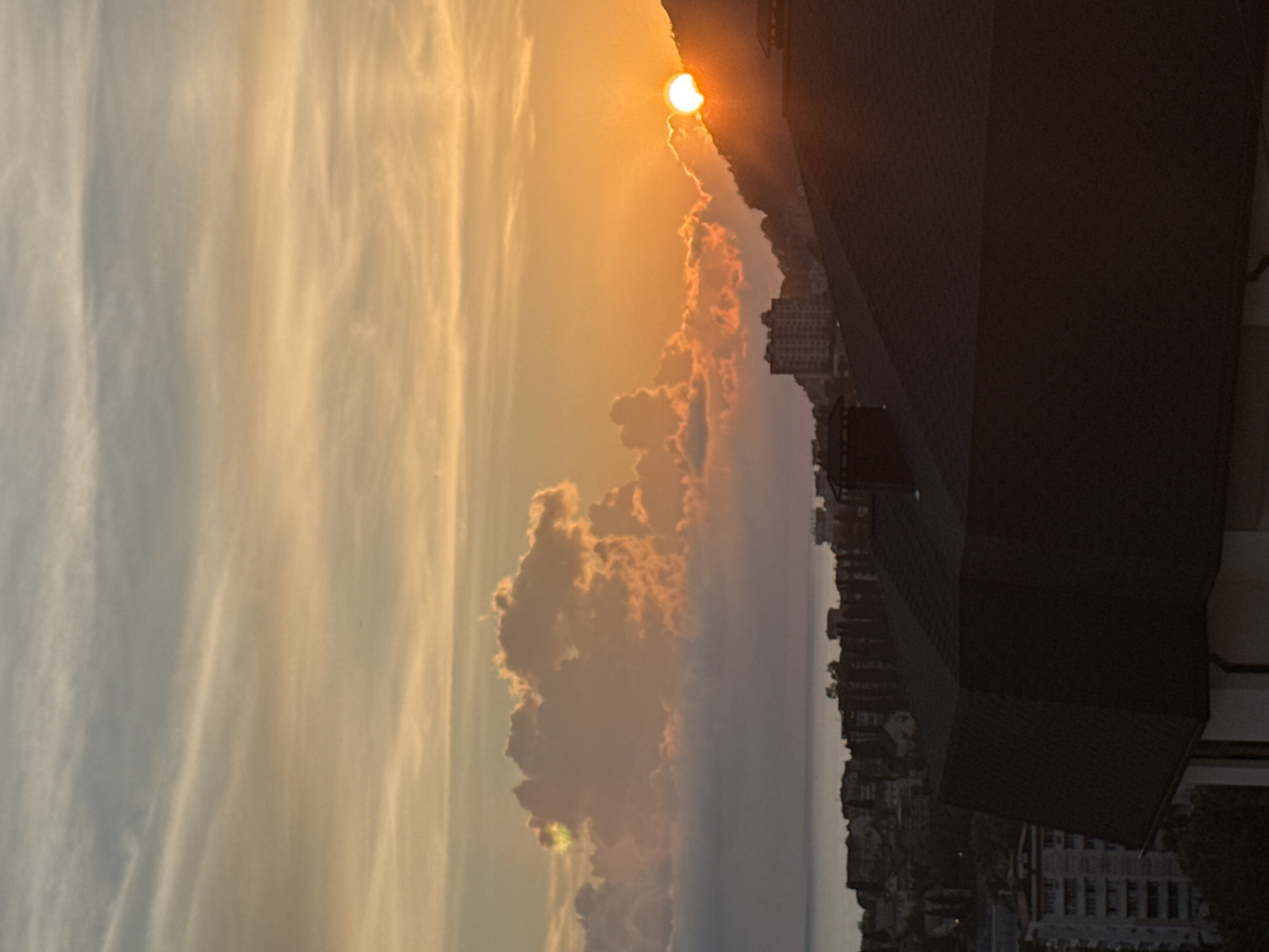 An outdoor, eye-level, vertical, medium shot captures a sunset over a city in Sochi, Russia. The sky is a gradient of pale blue, white, and orange, with wispy clouds illuminated by the setting sun. The sun itself is a bright orb partially obscured by a distant mountain range and clouds. In the foreground, the dark, angled roof of a building dominates the bottom of the frame. Behind it, a cityscape unfolds with buildings of varying heights and architectural styles, nestled against a hillside. The sea is visible in the background, with its surface calm and reflecting the colors of the sky. The overall atmosphere is serene and tranquil, with the golden hues of the sunset casting a warm glow over the scene.