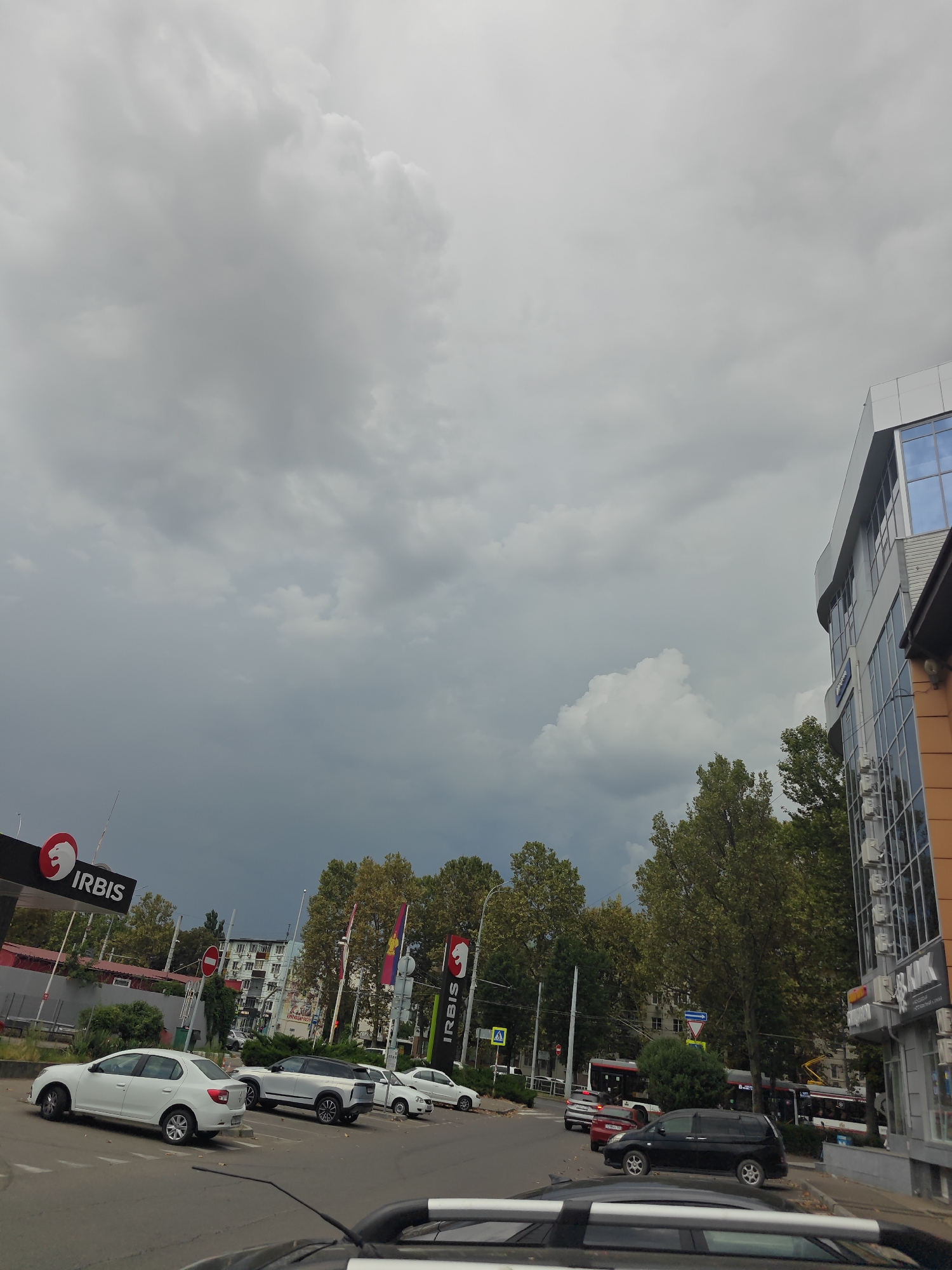 The image depicts a street scene in Krasnodar, Russia, likely on a cloudy day. In the foreground, several white cars are parked in a lot. Behind them, a gas station sign with "IRBIS" logo is visible. Further back, trees line the street, and a multi-story building with glass windows stands on the right side. A bus is also seen in the distance. The sky is overcast with dark clouds.