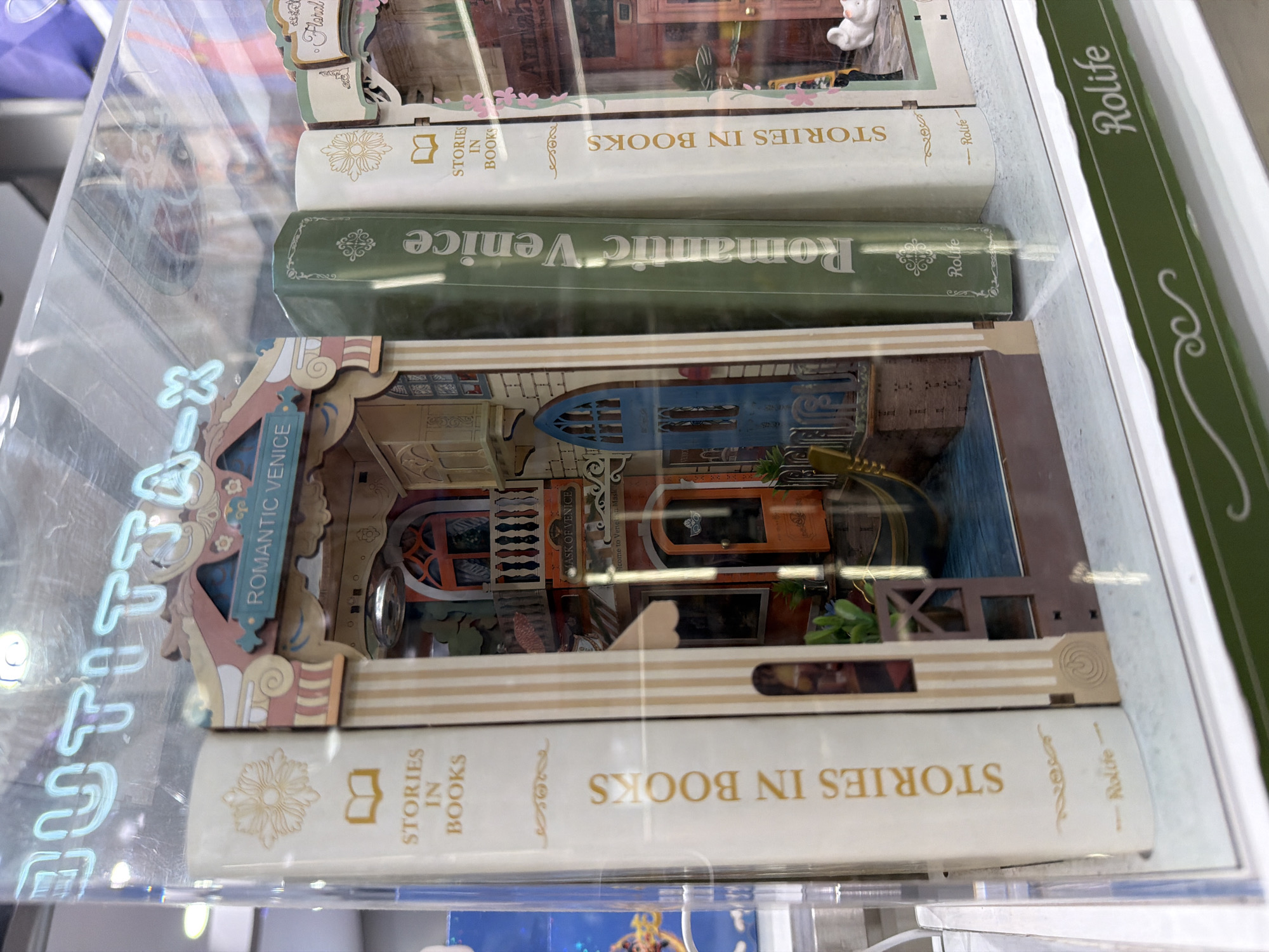 The image shows a display of miniature book-shaped dioramas. The central diorama depicts a scene inspired by Venice, with a balcony, a door, and a gondola. To the left of this diorama is a white book with gold lettering that reads "STORIES IN BOOKS" and a decorative element. To the right of the central diorama are two more book-shaped dioramas. The first is a green book with gold lettering reading "Romantic Venice." Next to it is a white book identical to the one on the left. The background is a transparent display case with a neon sign at the top that is partially visible and reads "BUTTITA-X". The scene appears to be an indoor retail display, possibly in a gift shop or a specialty store. There is no information in the image to suggest the location is in Milan, Italy.