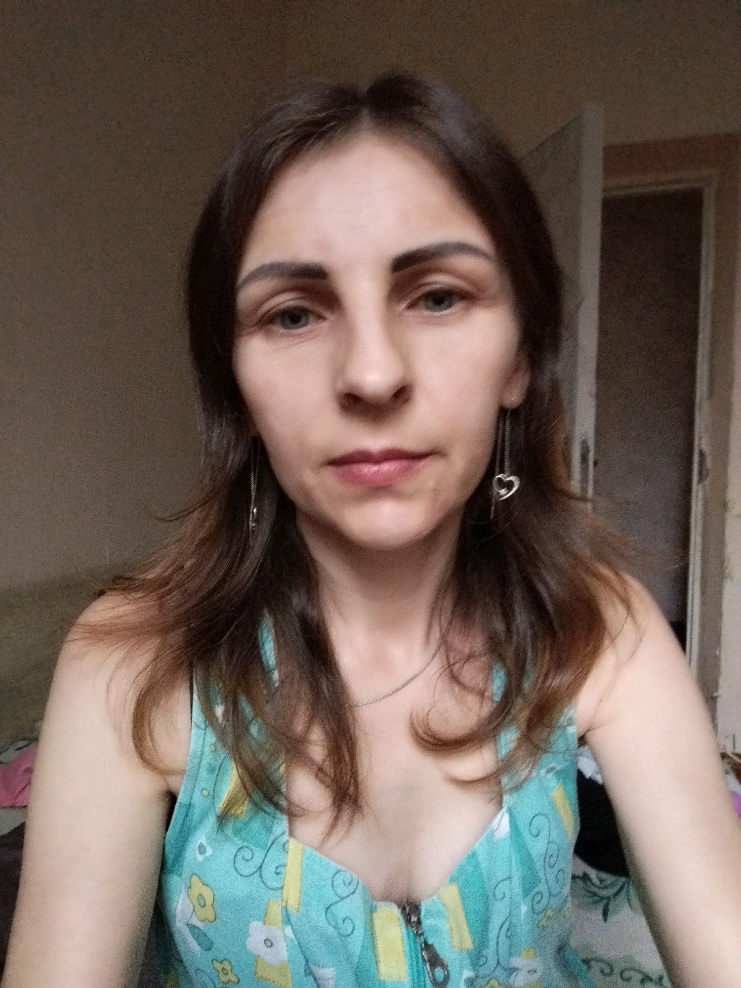 A medium shot from a slightly low angle shows a fair-skinned woman with dark brown hair and light blue eyes. She is looking directly at the camera. Her hair is shoulder-length and slightly wavy. She is wearing a teal, sleeveless dress with a yellow and white floral pattern. The dress has a zipper in the front. She is wearing a thin silver chain necklace and dangling earrings with heart-shaped charms. The background is a dimly lit room. To the left, a wall appears to be covered in textured wallpaper. To the right, a doorway is visible, with a door ajar, revealing a darker interior. The lighting is soft and appears to be coming from the front, casting subtle shadows. The overall impression is of a candid, indoor photograph. The image does not contain any identifiable landmarks or elements that would allow for pinpointing its location in Vinnytsia, Ukraine.