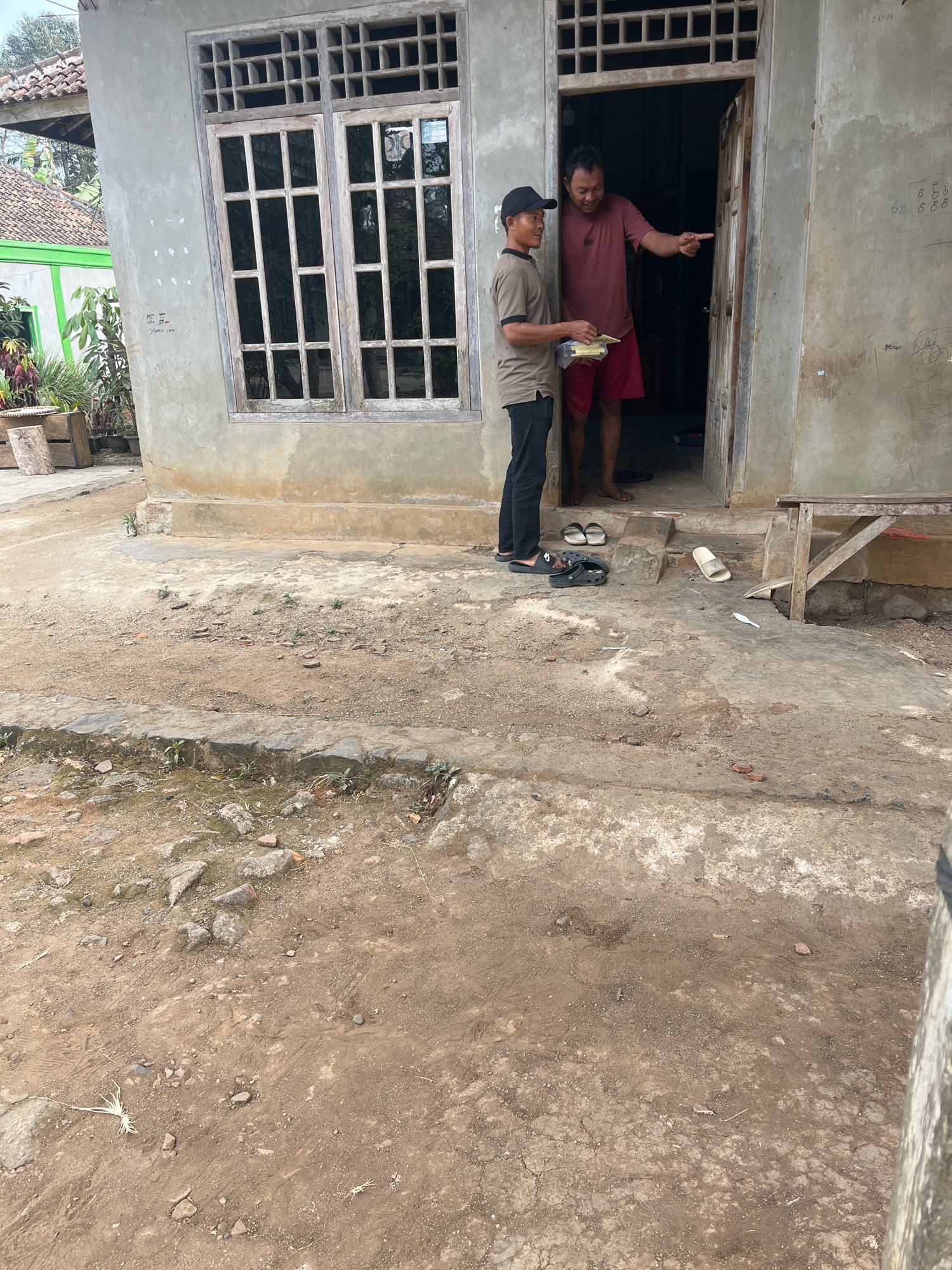 Two adult males are standing in the doorway of a simple, weathered concrete house with a wooden framed window. One man, wearing a dark baseball cap, a light brown t-shirt, and black pants, holds a rectangular object, possibly food, in his hands. He is facing a second man, who is taller and wearing a maroon t-shirt and red shorts. The second man is pointing with his right index finger towards the left, away from the camera, and appears to be speaking. The ground in front of the house is dirt and concrete, with scattered rocks and some dried vegetation. Several pairs of footwear are visible on the ground near the doorway, including black slides, black Crocs, and a white sandal. A wooden bench or table is positioned to the right of the doorway. The house itself has a textured concrete facade, and graffiti is visible on the wall to the right of the door. The background shows a tiled roof and some greenery, suggesting a residential area in Muara Dua, Indonesia, likely a rural or semi-rural setting.