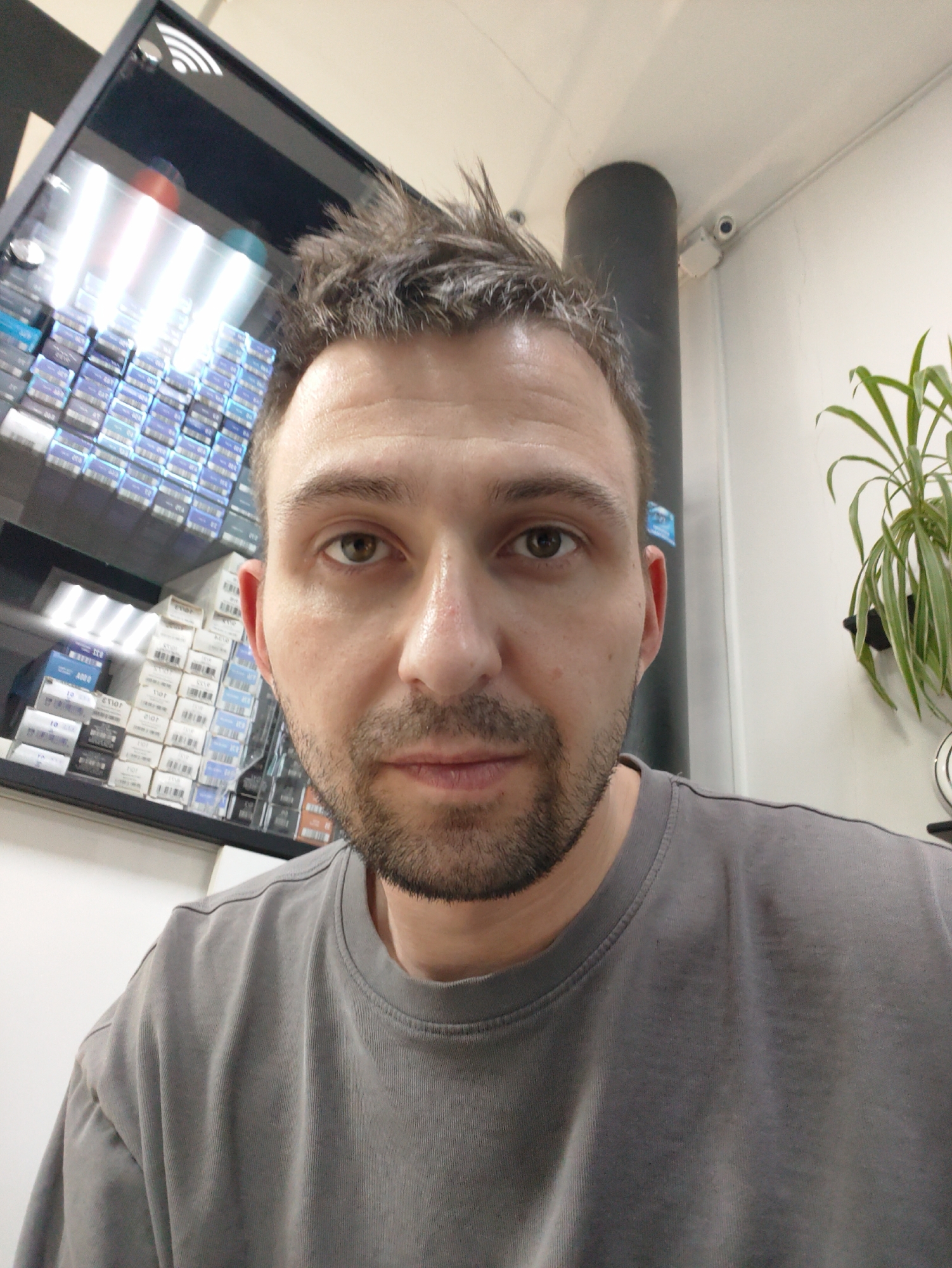 A man, likely in his 30s or early 40s, is centered in a close-up, selfie-style photograph. He has short, spiky brown hair and light stubble, with a neutral expression as he looks directly at the camera. His skin appears slightly reflective or oily, and he is wearing a plain grey t-shirt.
The background behind him suggests an indoor commercial environment. To the man's left, a glass display cabinet is filled with numerous small, rectangular boxes, predominantly white with blue accents. Reflections of bright interior lights are visible on the cabinet's surface, and a "Wi-Fi" symbol is present at the top left corner. Visible text and numbers on the boxes include "208," "02," "10," "1.0," and "1.5," some appearing mirrored.
To the man's right, a light-colored wall features a green potted plant and a dark grey vertical pipe or column. A small white security camera is mounted on the wall or ceiling near the column. The overall lighting is bright, indicating artificial indoor illumination. The man is not engaged in any specific activity other than posing for the photograph.