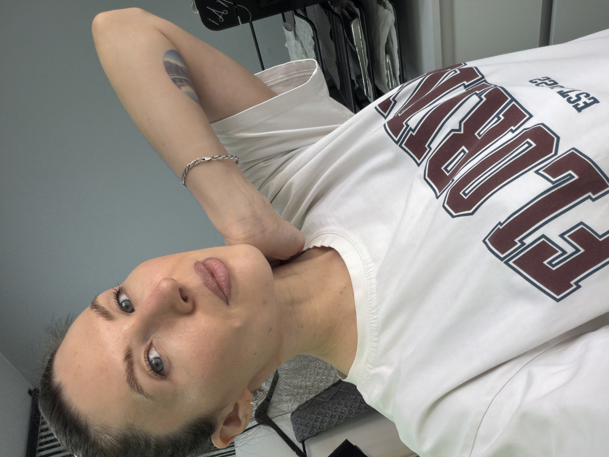 A woman is pictured indoors, possibly in a private residence. She is wearing a white t-shirt with a collegiate-style maroon and blue logo on the front. She has fair skin, blue eyes, and light brown hair. She is wearing a silver bracelet on her left wrist and a silver earring on her left ear. There is a tattoo on her left arm. Behind her is a clothes rack and a light gray wall. The city and country are not discernible from this image.