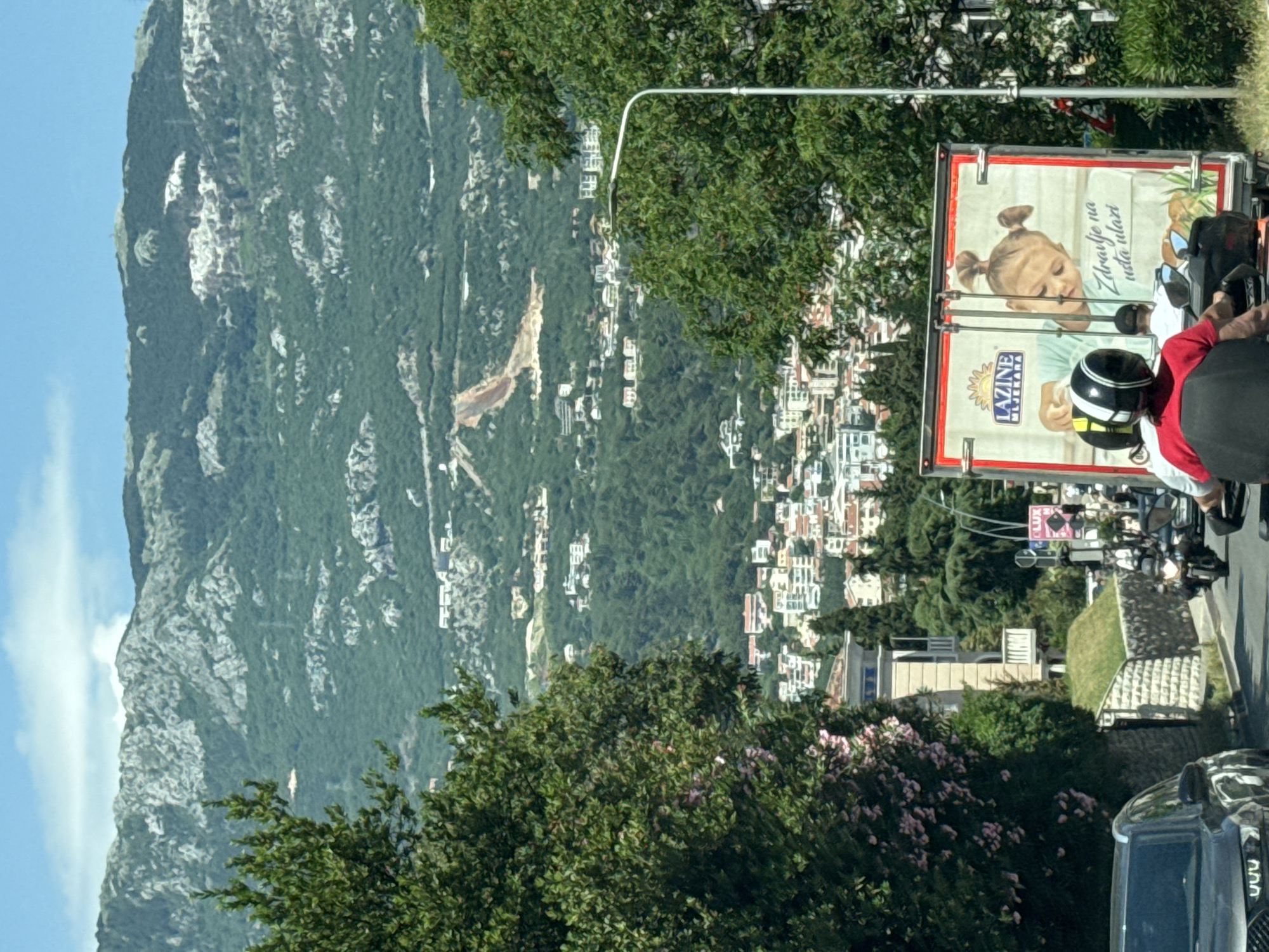 The image depicts a landscape scene in Budva, Montenegro. A large, mountainous terrain dominates the left side of the frame, with a mix of rocky terrain and forested areas. On the right side of the image, a residential area is visible, with closely built houses nestled among dense greenery. In the foreground, a tree with pink flowers is visible. A motorcycle is being driven on the road with a Lazaine Njelakara billboard attached to the back. A black car is partially visible at the bottom of the frame.