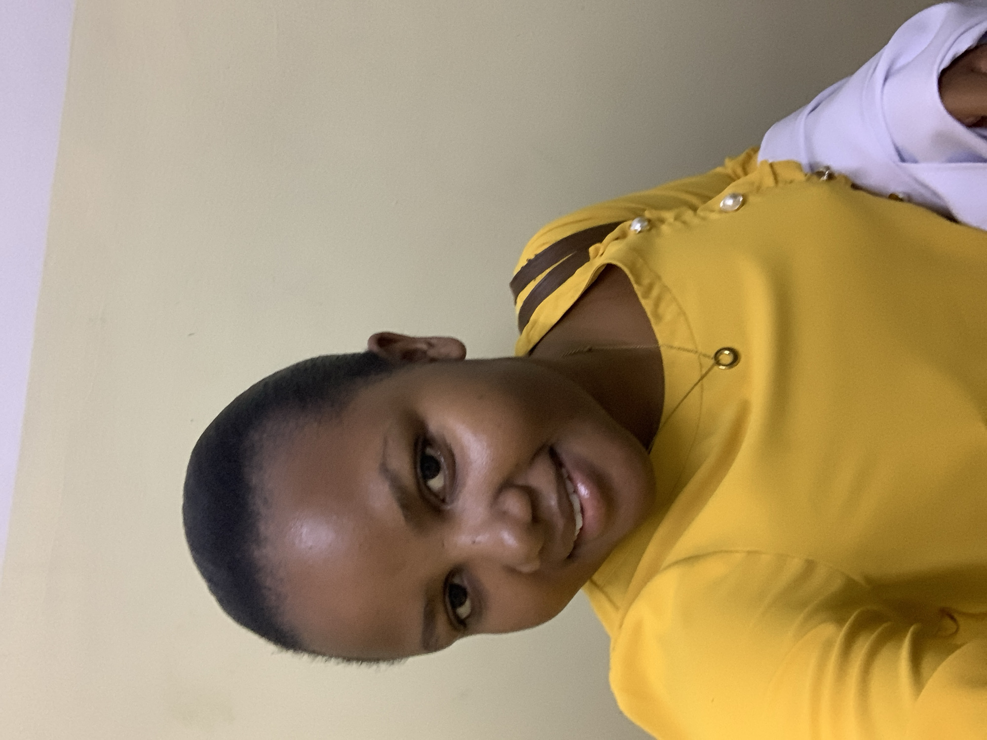 A woman is posing for a picture indoors. She has short, dark hair and is wearing a yellow shirt with decorative buttons. Part of another person wearing white clothing is visible in the upper right corner of the frame. The background is a plain, light-colored wall. No information indicates that the location is Dar es-Salaam, Tanzania.