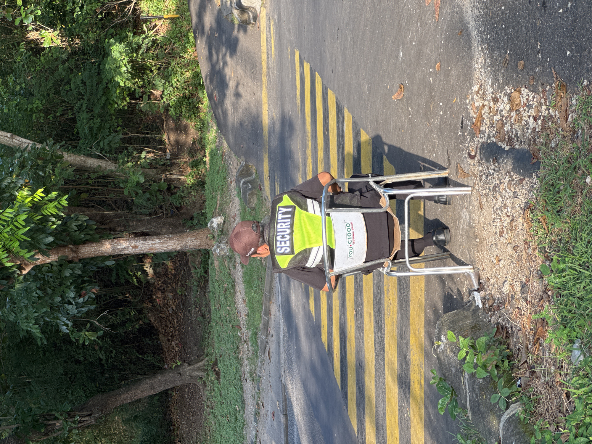 A security guard sits in a chair, on a road with yellow stripes, in Kuta Selatan, Indonesia. The guard wears a brown cap, a black shirt, and a yellow vest that reads "SECURITY". The chair is silver. A sign for "You-C1000 Isotonic Drink" is visible on the chair. To the left is a wooded area, and to the right is grass.
