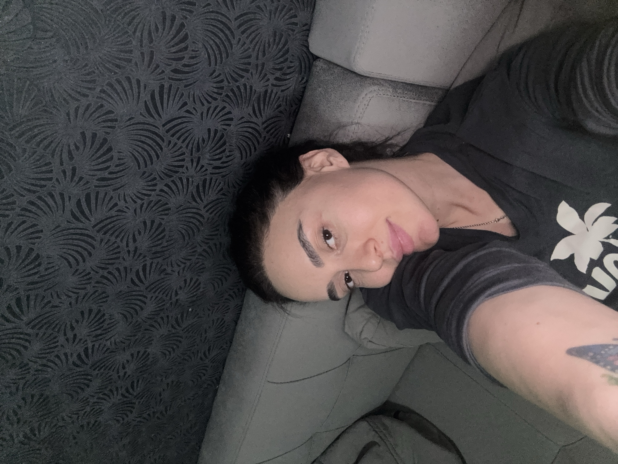 A woman with dark hair is lying on a gray sofa. She is wearing a dark gray t-shirt with a white floral design. She has a tattoo on her arm. The wall behind the sofa has a dark gray patterned wallpaper. The location appears to be indoors, but there is no evidence to suggest it is in Tbilisi, Georgia.