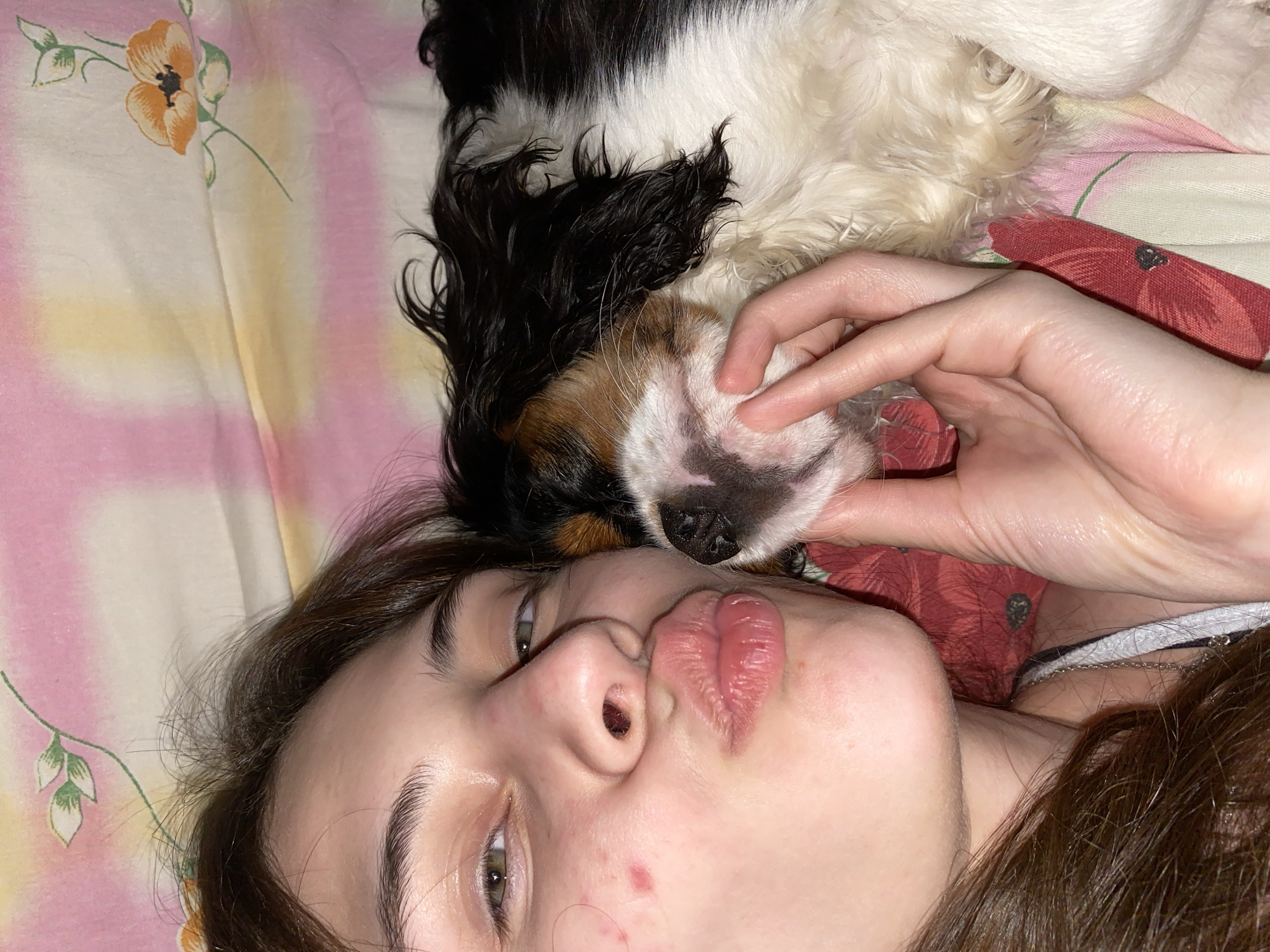 An intimate overhead shot, rotated on its side, captures a young woman and her small dog lying together in a domestic setting, likely a bedroom in Votkinsk, Russia. The woman, with long dark hair and fair skin, looks towards the camera with green eyes, her lips playfully pursed into a "kissy face." Her left hand gently cradles the chin of a tri-color dog, identifiable by its black, white, and tan fur, possibly a Cavalier King Charles Spaniel, whose head rests beside hers.
The subjects are positioned on a soft surface, likely a bed, covered by a patterned sheet or duvet. The bedding displays a gentle tie-dye effect with shades of pink, yellow, and white, adorned with illustrative orange poppy flowers and green leaves. A glimpse of red patterned fabric, possibly clothing or another blanket, is visible beneath the woman's arm and behind the dog. A delicate silver chain is also visible around the woman's neck area. The scene radiates affection and a relaxed, playful mood. No text is visible in the image.