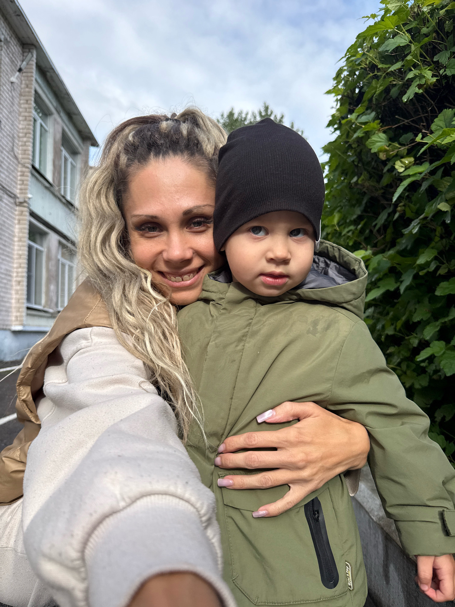 In the city of Rusanovo, Russia, a woman with blonde, wavy hair pulled into a ponytail smiles at the camera. She is wearing a tan jacket over a cream-colored hoodie. She is embracing a young boy who is wearing a black beanie and a green hooded jacket. The boy looks forward with a neutral expression. They are positioned in front of a building with light-colored brick and several windows. To the right of the subjects, there is a lush green hedge. The sky above is overcast.
