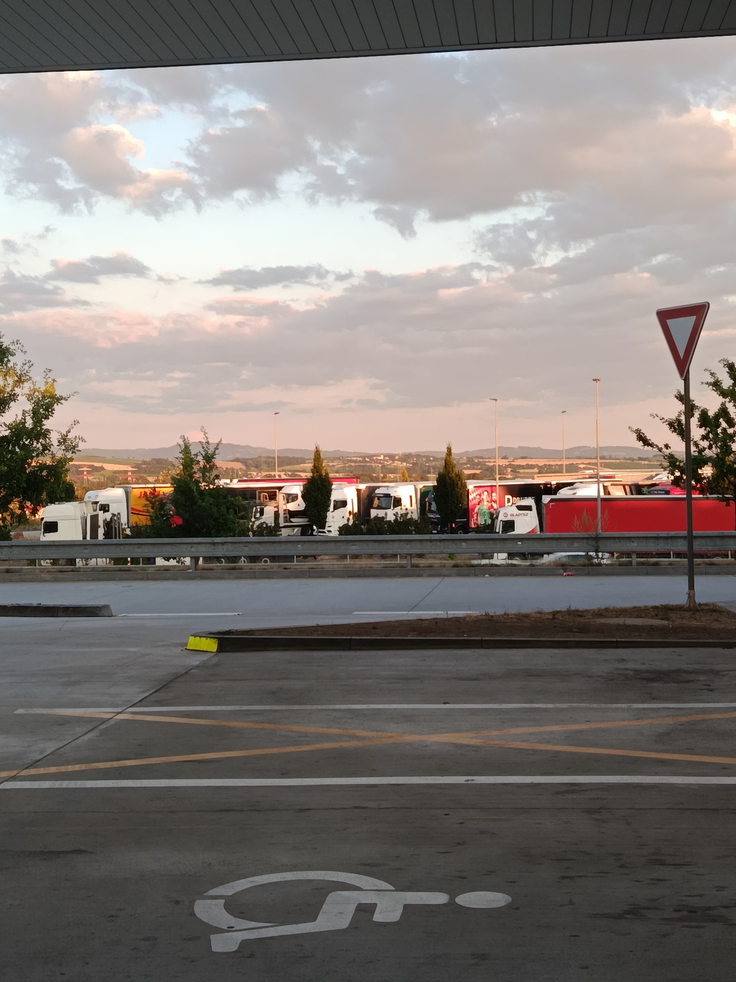 The image captures a parking area for semi-trailer trucks, likely a rest stop or depot, under a soft, cloudy sky at dusk. Several white and red trucks are parked in a row, some with trailers, indicating a commercial trucking operation. The foreground shows a paved surface with yellow and white lines, designating parking spaces, and a white symbol for disabled parking is visible on the ground. A red and white triangular yield sign is present on the right side of the image, along with a tree. In the background, rolling hills and distant structures suggest a rural or semi-rural setting. The overall impression is one of a functional transportation hub at the end of the day. The specific location in Osek nad Bečvou, Czechia, is not visually evident from the image alone but would be external knowledge.