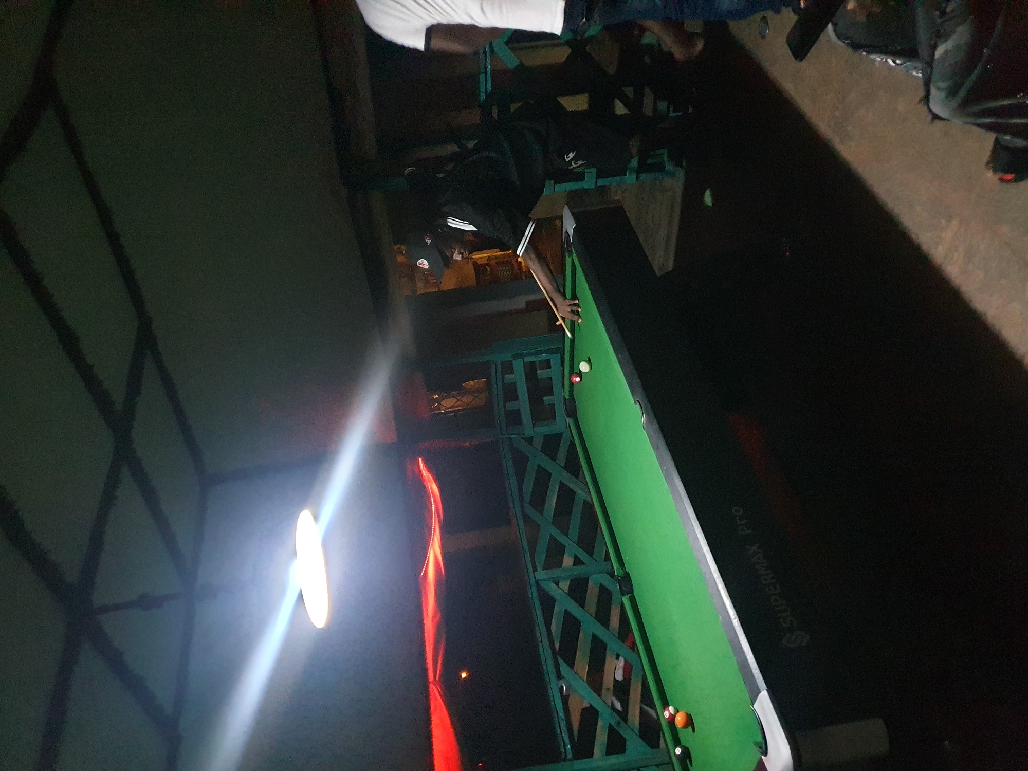 A nighttime image taken in Lokoja, Nigeria, shows a man in dark clothing and a cap playing pool. He is positioned over a green pool table, cue in hand, aiming at the balls. Parts of other individuals are visible at the top of the frame. The pool table, identified by the "Supermax Pro" logo, is situated on what appears to be a balcony or elevated platform with a green wooden railing. A bright light source casts harsh shadows on the structure to the left. A red curtain or fabric hangs in the background. The environment suggests a casual, possibly recreational, setting.