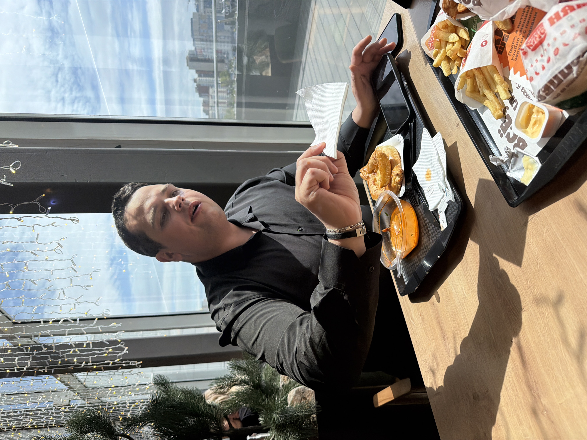 The image shows a man in a black long-sleeved shirt seated at a wooden table in what appears to be a fast-food restaurant. He is holding a white napkin in his right hand and looking towards the camera. Next to him is a black tray containing a croissant, a small container of sauce, and a smartphone. On the table, there is another black tray with fast food, including french fries, burgers wrapped in paper with Cyrillic text, and a container of sauce. Behind the man is a large window overlooking an urban landscape with multiple buildings and a cloudy sky. There are some decorative light strings visible near the window. Based on the Cyrillic text on the fast-food packaging, the location is likely in Russia. Based on the available knowledge, the city is Krasnoyarsk.