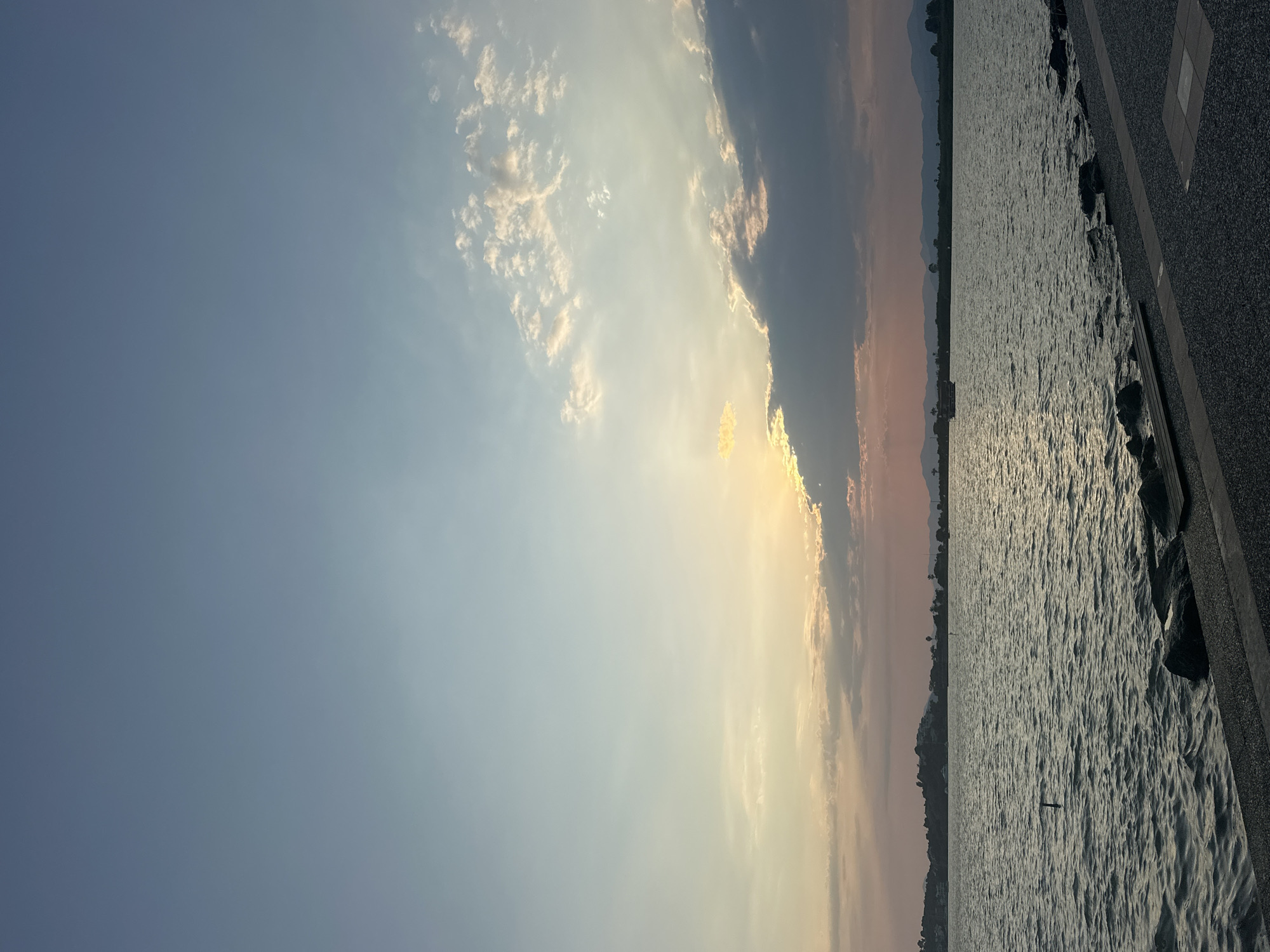 The image depicts a serene waterscape at dusk, likely along the coast of Fethiye, Turkey. The sky transitions from a soft, pale blue at the top to hues of pink and orange near the horizon, where the sun is setting behind a bank of dark clouds. Wisps of lighter clouds catch the fading sunlight, creating a gentle, diffused glow.
The body of water, possibly a bay or lagoon, is calm with gentle ripples reflecting the sky's colors. In the distance, a silhouetted landmass with rolling hills and vegetation stretches across the horizon. A few scattered trees are visible on the shoreline, and the faint outline of mountains can be seen further inland. A small boat is present on the water, contributing to the peaceful atmosphere.
In the foreground, a paved walkway or embankment lines the water's edge. The walkway is composed of dark gravel and features distinct square tiling in places. Rocks of varying sizes are placed along the edge of the water, some partially submerged. A wooden bench or platform is also visible on the embankment. There are no people or animals in the frame. The scene is characterized by natural beauty and tranquility.