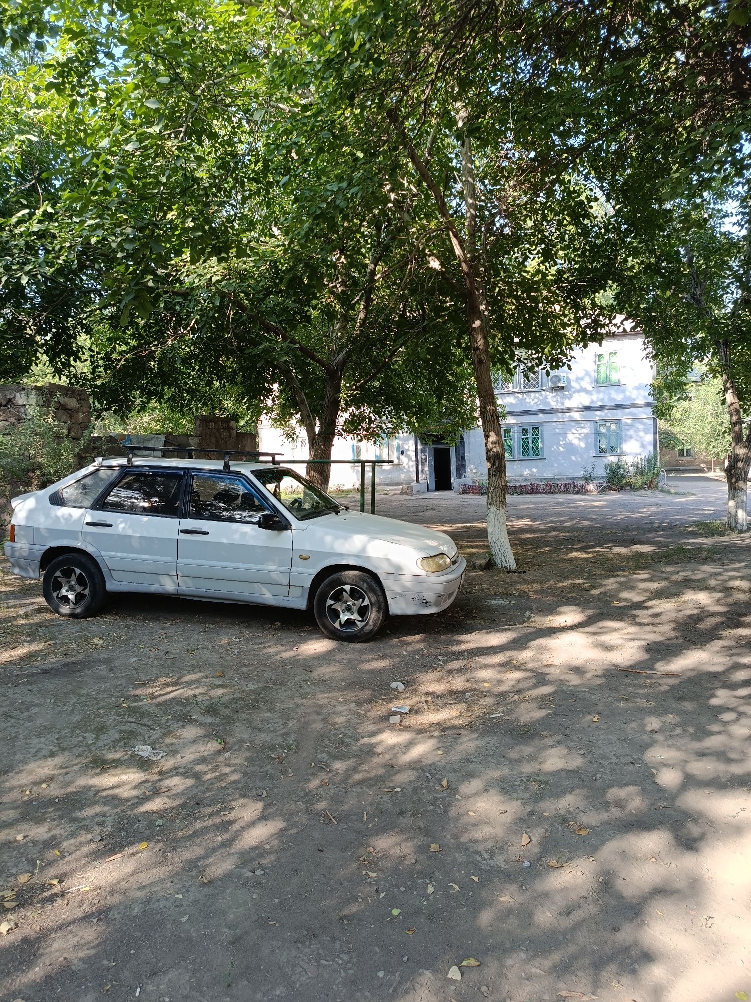 A white, five-door hatchback car with a roof rack is parked on a dirt lot. The car is angled to the left, with its front facing the right side of the frame. The car appears to be old and slightly weathered, with some visible rust and dirt. In the background, there is a light-colored, two-story building with green-trimmed windows and a faint cross-hatch pattern on some of them. The building is partially obscured by lush green trees, with sunlight dappling through the leaves, casting shadows on the ground. The ground is uneven and covered in dirt, with scattered leaves and debris. A stone wall is visible to the left of the car. The scene suggests a residential or semi-rural setting, possibly in a warm climate given the foliage.