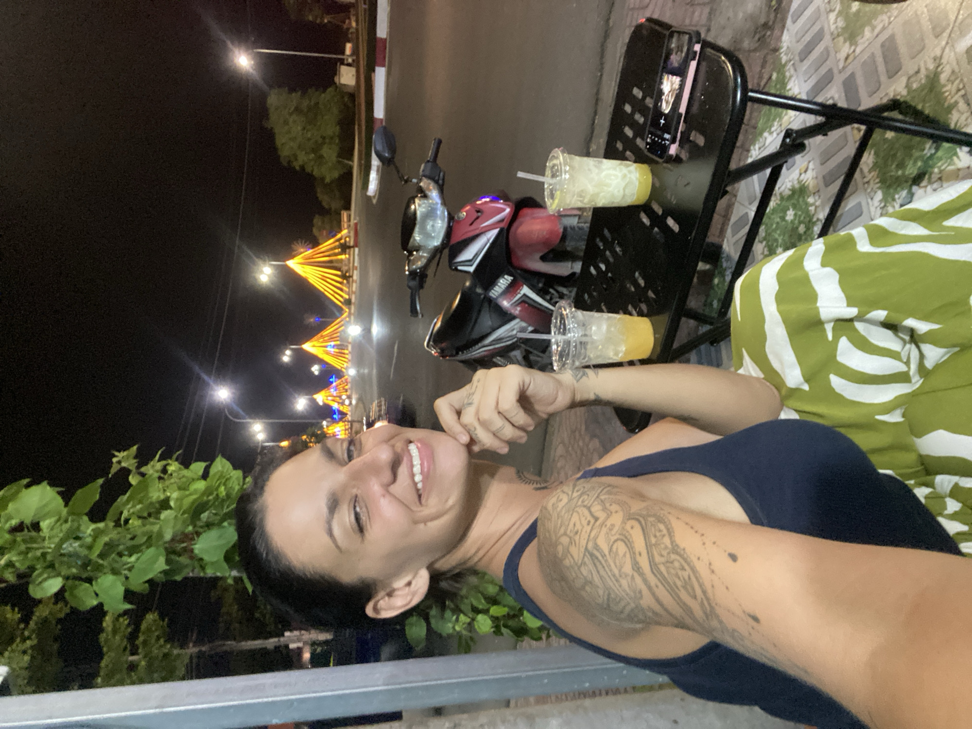 A woman is captured in a selfie at night, seated at a black metal table on a sidewalk. She has dark hair pulled back and is smiling, her head tilted slightly. Her left arm, adorned with intricate tattoos, rests on the table, and she is wearing a dark blue tank top and a green and white patterned skirt. Two clear plastic cups with straws, one containing a yellow beverage and the other a light-colored beverage, sit on the table. A smartphone, with a pink case and a preview of a photo visible on the screen, is also on the table. Behind her, a red and black Yamaha motorcycle is parked. The background reveals a street scene at night with illuminated decorative pyramid-shaped structures in yellow and orange lights, streetlights, and trees. The environment suggests an urban setting, specifically Phu Thuy Ward, Vietnam, indicated by the presence of the motorcycle and the style of the decorative lighting.