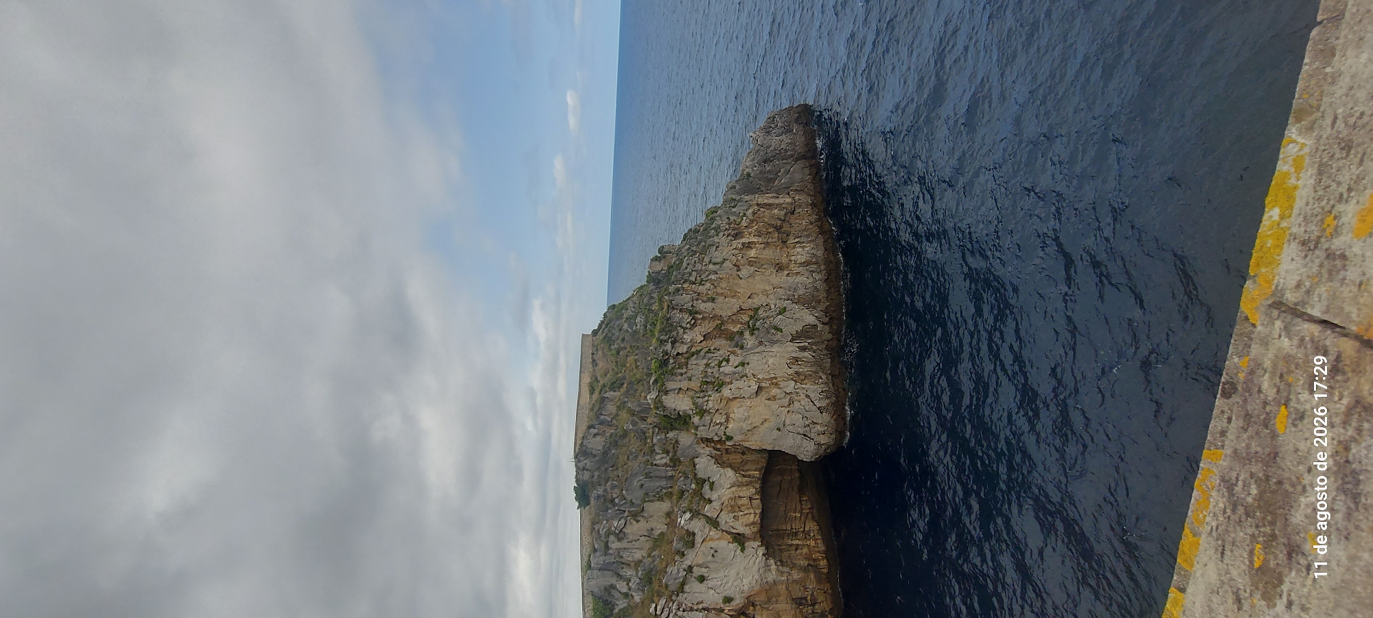 The image shows a rocky cliff overlooking a dark blue sea under a cloudy sky. A cave is visible within the cliff face. The top of the cliff is flat and appears to be a historical structure, possibly a fortification or ruins, with low stone walls. Green vegetation is scattered across the cliff. In the foreground, a concrete ledge with yellow markings is visible. A timestamp "11 de agosto de 2026 17:29" is superimposed on the bottom left of the image. The scene depicts a natural coastal landscape with historical elements. While the image does not explicitly show Castro-Urdiales, the description of the landscape aligns with coastal areas of Northern Spain, which could potentially be Castro-Urdiales.