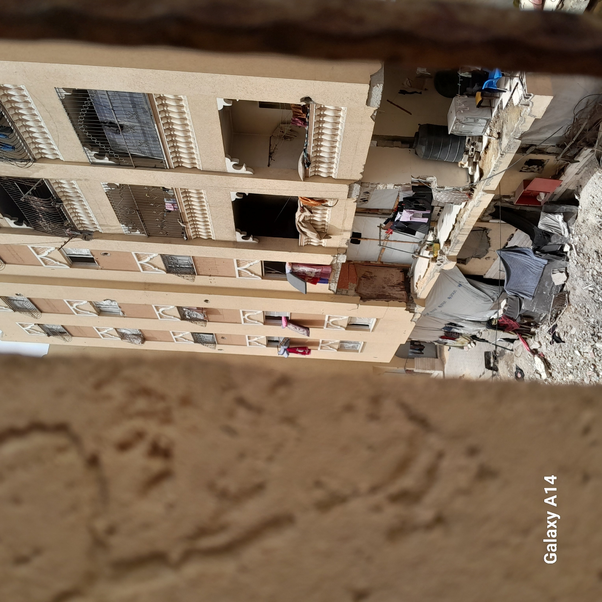The image provides a perspective looking down from an elevated position, focusing on a multi-story building and a cluttered ground or lower roof area adjacent to it.
The main subject is a light-colored (beige/tan) multi-story building, likely residential, dominating the left and center of the frame. It features numerous windows, many of which are fitted with ornate metal grates. Some windows and balconies have decorative white architectural elements. Clothes are visibly hanging to dry from several windows and open balcony areas on different floors. One large, dark, open section is present on an upper floor, appearing as a recessed balcony or an empty opening.
To the right of the building, a ground-level or lower roof area appears quite disheveled and cluttered. This space contains two large, dark, cylindrical tanks (possibly water storage tanks), a large white appliance resembling a washing machine, and a blue plastic chair. Various tarps and sheets are spread across this area; one prominent blue and white tarp clearly shows the "UNICEF" logo. Clotheslines with laundry are also strung across this space. The ground itself is uneven and strewn with debris, rocks, and dirt, suggesting either a state of significant disrepair, ongoing construction, or an informal/makeshift living or storage area.
The implied activity is domestic, specifically laundry, as indicated by the numerous clothes hanging out to dry. No people are distinctly visible. The lighting suggests it is daytime. The overall scene depicts an urban or semi-urban environment, potentially in an area with informal settlements or buildings in varying states of maintenance. The text "Galaxy A14" is visible in the bottom right corner of the image (when oriented horizontally), indicating the device used to capture the photo.