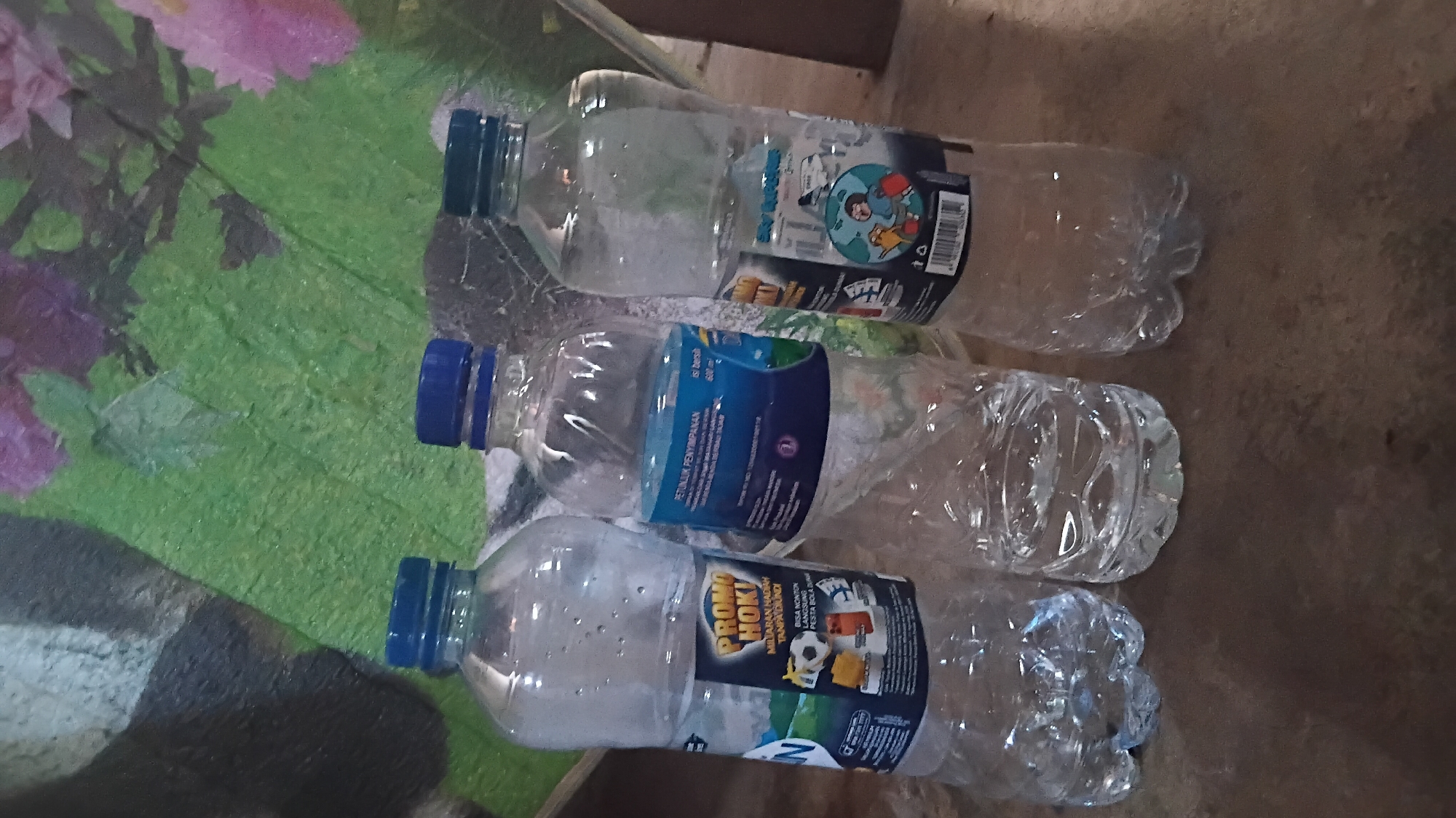 Three empty plastic water bottles with blue caps are arranged in a line on a textured surface. The leftmost bottle has a yellow and black label that reads "PROMO HOKI" and "MILIARAN HADIAH TANPA DIUNDI". The middle bottle has a blue and white label with Indonesian text. The rightmost bottle has a dark label with an illustration of a person holding a globe. The background is a green textured surface with purple and pink floral elements. The image appears to be taken indoors, possibly in a residential setting in the city of Prabumulih, Indonesia, given the context of the labels. The lighting is dim, casting shadows on the bottles.