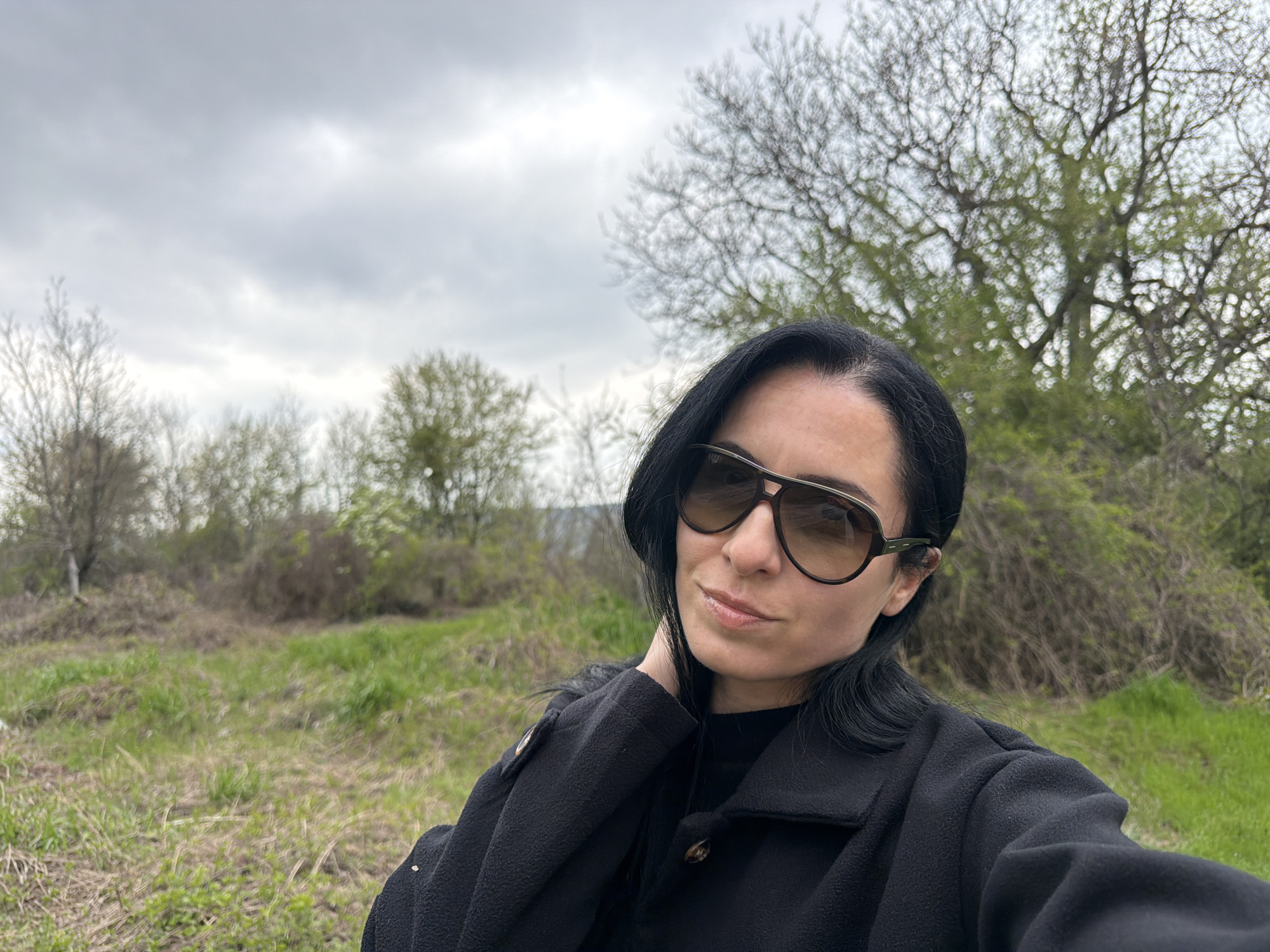 A woman wearing sunglasses and a black coat stands in a grassy field with sparse trees under a cloudy sky. The location appears to be in a rural area on the outskirts of a city, based on the distant vegetation and sky conditions. Given the prompt, this would be in the city of Pleven, Bulgaria, and the surrounding area.