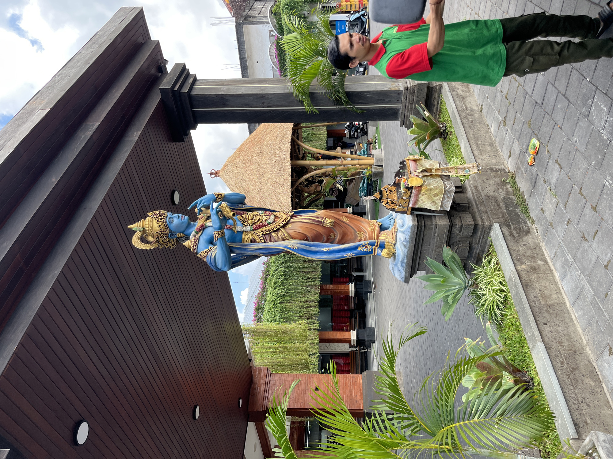 The image shows a statue of a blue-skinned deity, likely from Hindu mythology, standing in an outdoor courtyard. The statue is elaborately decorated with gold accents and wears a crown. It stands on a stone pedestal amidst lush greenery and tropical plants. In the background, a traditional thatched-roof structure is visible. A man in a green and red shirt and dark pants is standing to the right of the statue, looking away from the camera. The scene appears to be set during the daytime, with diffused sunlight suggesting either overcast skies or a partially shaded area. The paving stones and architecture suggest a warm climate, and the location context of Kuta, Indonesia, confirms a tropical setting. There is no visible text in the image. The overall atmosphere is one of cultural significance and serene outdoor ambiance.