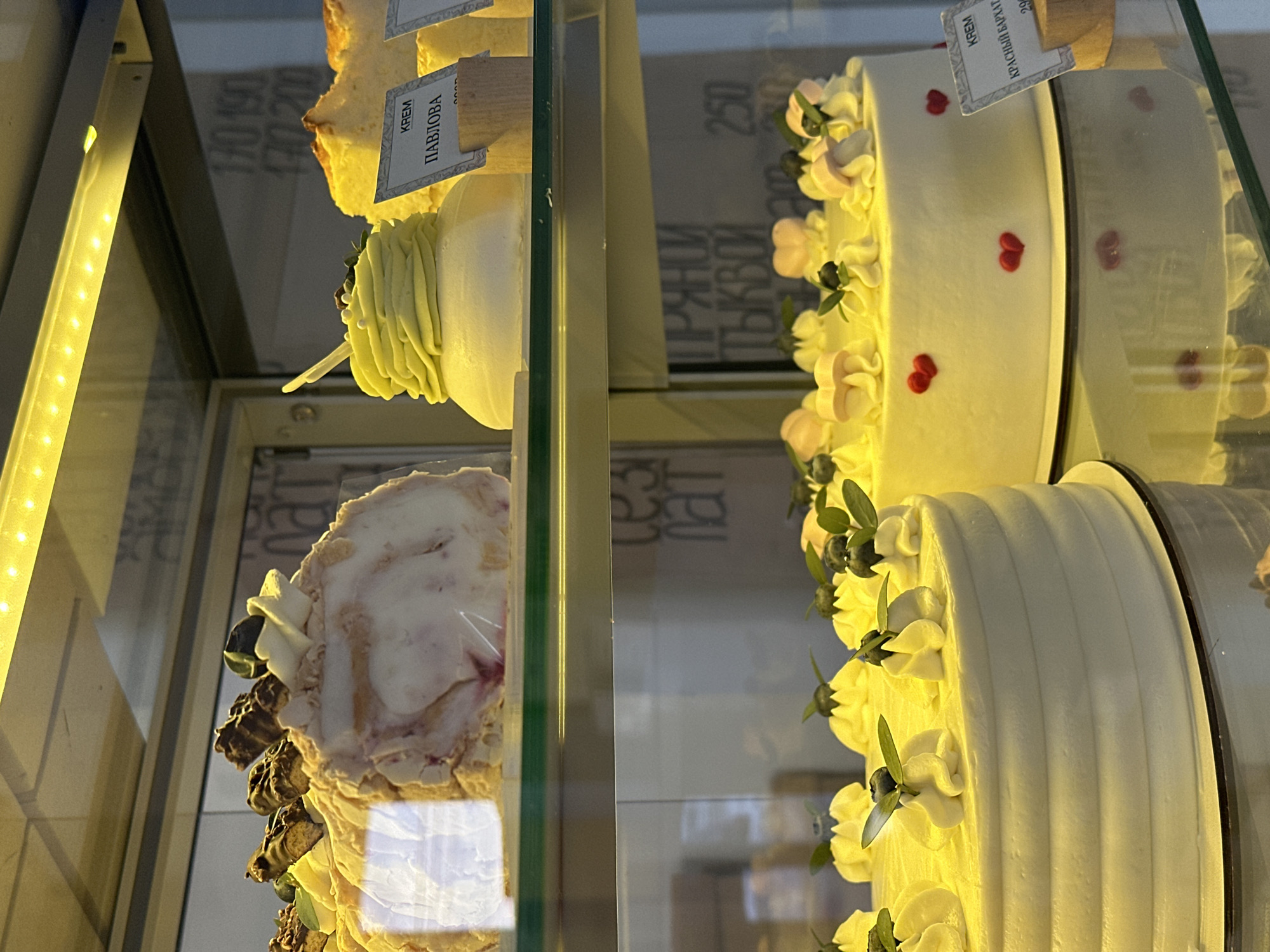 The image displays a brightly lit glass pastry display case in a confectionery located in Votkinsk, Russia. The case contains two shelves of assorted desserts.
On the upper shelf, from left to right:
* A large, elongated, light-colored meringue or cream roll, garnished with dark brown confectionery pieces and small green accents.
* An individual serving dessert with a white base topped with light green piped cream, accented with small white spheres and a dark berry or leaf. A small wooden sign in front labels it "КРЕМ ПАВЛОВА" (Cream Pavlova).
* Portions of a yellow-colored cake, likely sponge cake.
On the lower shelf, from left to right:
* A round, multi-layered cake with smooth white frosting featuring horizontal ridges on its side. The top is decorated with numerous yellow cream swirls, each adorned with a blueberry and a small green leaf.
* A large, round cake with smooth white frosting, decorated with several small red heart shapes applied to its side. The top features similar yellow cream swirls, blueberries, green leaves, and light pink mushroom-shaped confectionery decorations. A small wooden sign labels this cake "КРЕМ КРАСНЫЙ БАРХАТ" (Cream Red Velvet) with a visible price starting at "29".
The display case is illuminated by an overhead yellow LED strip. Reflections on the glass surfaces reveal fragmented Cyrillic text and numbers, including "170 190", "170 200", "ТЕЗЫ ПАТ", and "ПРЯНОЙ ТЫКВОЙ 250" (Spiced Pumpkin 250), indicating additional products or pricing information beyond the immediate scope of the image.