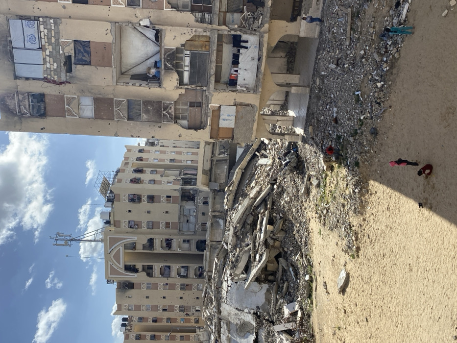 The image depicts an urban environment in Khan Yunis, Palestinian Territory, characterized by extensive structural damage.
In the foreground, a sandy patch of ground contains scattered debris and intermittent small patches of vegetation. Three individuals, identified as children, are present on this sandy ground: two are kneeling or sitting, and one stands upright. One child wears a pink hooded garment, another a light blue/green garment.
The midground is dominated by a large mound of pulverized concrete and rebar rubble, indicative of a collapsed multi-story structure. This debris pile is irregularly shaped and occupies a significant portion of the central-left frame.
To the right, a multi-story residential building, light beige in color, exhibits severe damage. Multiple sections of its facade are absent, exposing internal rooms. Windows are either missing, shattered, or covered with makeshift materials such as cardboard, tarpaulins, or metal sheeting. On an upper floor, clothes are visible hanging on a line within an exposed interior section. The building's exterior surfaces are marked with numerous small, circular impact holes. At ground level, the building features a series of arched pedestrian passages. An adult male, wearing a blue shirt, stands near the base of this damaged building.
In the background, another multi-story residential building, similar in architectural style, displays damage including numerous small impact holes on its facade and broken windows. This building features a distinctive decorative arch on its upper-middle section. Its rooftop is equipped with multiple black water tanks, solar panels, and a prominent communications tower supporting several antennas.
The sky is partly cloudy with patches of blue visible. The scene is illuminated by natural daylight, casting shadows from the structures.