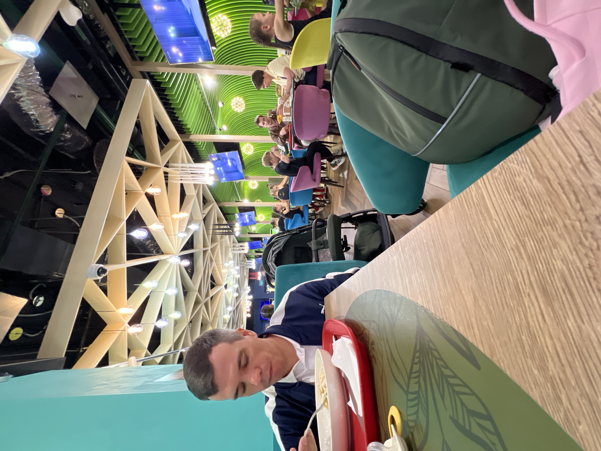 The image depicts a casual dining setting within a modern establishment. In the foreground, a man with short brown hair is seated at a table, wearing a white polo shirt under a dark blue and white jacket. He is in the process of eating from a white bowl with a fork. A red tray holding the bowl and a white napkin is in front of him. The table has a wooden surface and a green placemat with a leaf pattern.
The background reveals other patrons and the interior of the restaurant. Several people are seated at various tables, engaged in eating and conversation. The restaurant features vibrant decor with bright green wavy panels on the walls and ceiling. Numerous light fixtures, some resembling orbs and others elongated, are visible. Large screens are mounted on the green wall. The seating includes a variety of colorful chairs: teal, purple, and yellow. A baby stroller is parked to the right of the man in the foreground. A large green duffel bag is placed on a teal chair in the mid-ground. The overall impression is of a lively and contemporary eatery. The location is identified as a restaurant in Moscow, Russia.