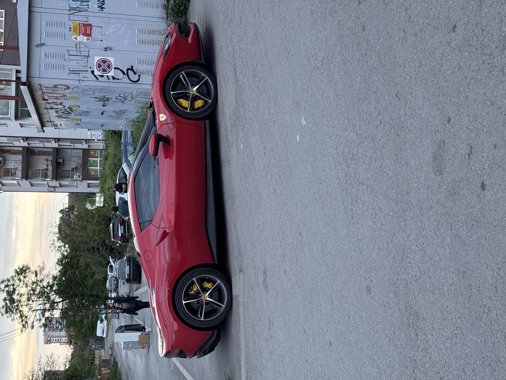 The image shows a red Ferrari parked on the street in Sofia, Bulgaria. The car is parked next to an electrical box with graffiti on it, and a multi-story building is in the background. A group of people stands behind the car near other parked vehicles. The street is grey asphalt. The time appears to be daytime.