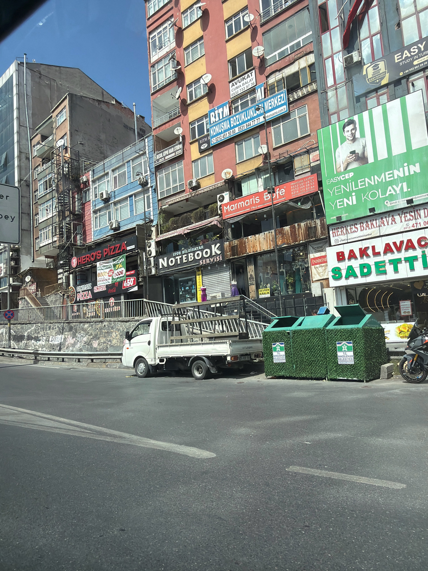 The image depicts a street scene in Bahçelievler, Turkey, on a clear, sunny day. In the foreground, a grey asphalt road occupies the bottom portion. A white truck with an open bed is parked on the right side of the road, facing left. Behind the truck, two green, hedge-covered recycling bins with official seals are placed. A motorcycle is parked to the right of the bins.
In the mid-ground, a stone retaining wall with graffiti runs horizontally. Above the wall, a narrow street or walkway with stairs leads up to various businesses. A white truck is visible here as well.
The background is dominated by a dense urban landscape of multi-story buildings. The buildings exhibit a variety of architectural styles and colors, including red, yellow, and blue facades. Several signs are visible on the buildings, advertising businesses such as "HERO'S PIZZA," "Marmaris Büfe," and "NOTEBOOK SERVİSİ." Other signs are in Turkish and appear to be for legal services and other commercial establishments. Satellite dishes are mounted on the exterior walls of some buildings. The overall impression is a bustling commercial and residential area.