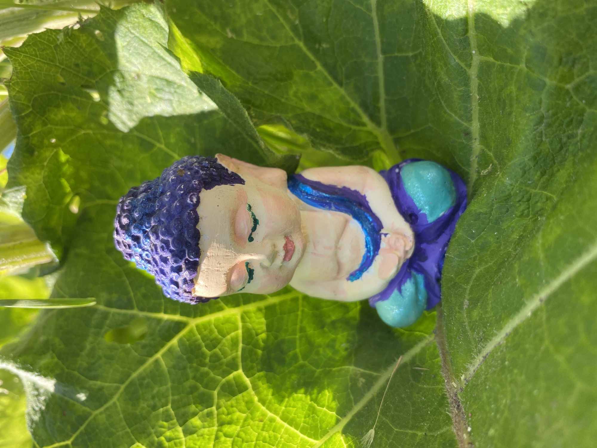 A small, colorful figurine of a meditating Buddha rests on a large, green leaf. The figurine is predominantly purple and teal, with a light-colored face and rosy cheeks. Its eyes are closed, and its hands are clasped in its lap. The leaf is vibrant green with visible veins and appears to be part of a larger plant, possibly a sunflower, given the surrounding foliage. The scene is illuminated by bright sunlight, casting shadows and highlighting the textures of the leaf and the figurine. The location is identified as Zelenodol'sk, Russia.