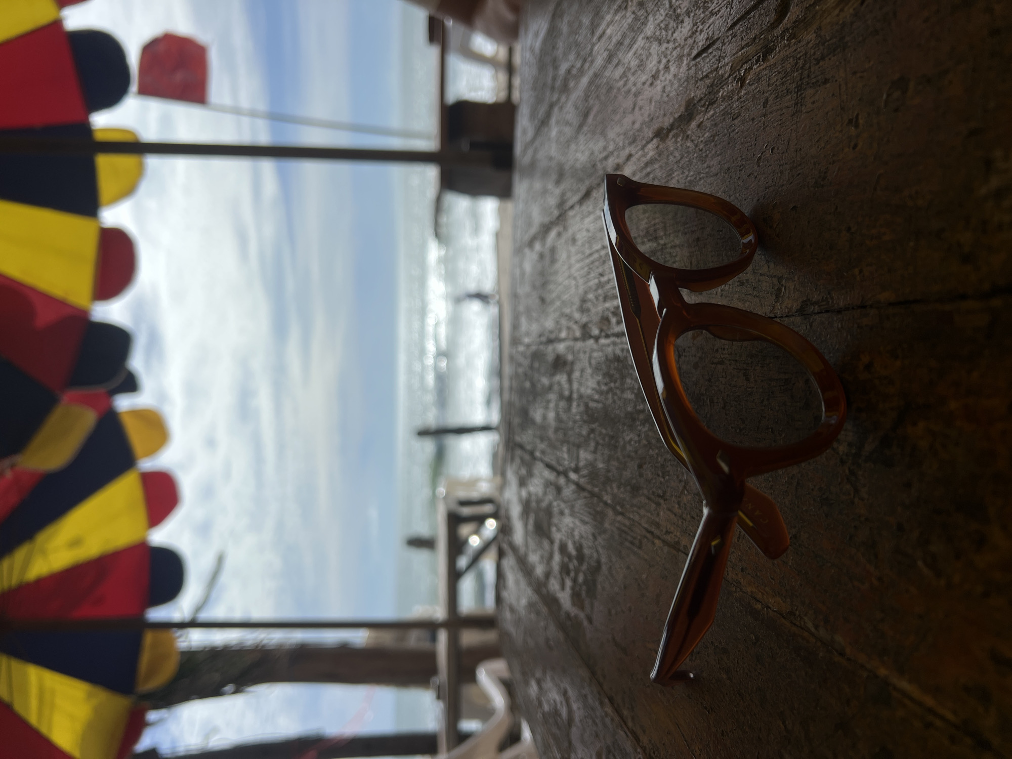 A pair of brown, thick-framed eyeglasses with circular lenses rests on a dark, weathered wooden table, sharply focused in the foreground. The table surface exhibits distinct grain and minor imperfections, possibly indicating dampness. Beyond the table, the scene gradually blurs into the background. A beachfront structure with wooden posts and crossbeams is visible, with faint outlines of white plastic chairs and other tables. In the mid-distance, an expanse of ocean shimmers under a bright sky, reflecting sunlight off the water. The horizon line separates the ocean from a partly cloudy sky. Several indistinct, blurry human figures are discernible near the water's edge. Overhead, partially framed, are segments of large beach umbrellas displaying alternating red, yellow, and dark (likely blue or black) colors. A red flag, possibly indicating a warning or designated area, is visible fluttering in the far right background, against the sky. The overall setting appears to be an outdoor, shaded area at a beachside location in Unknown, Unknown.