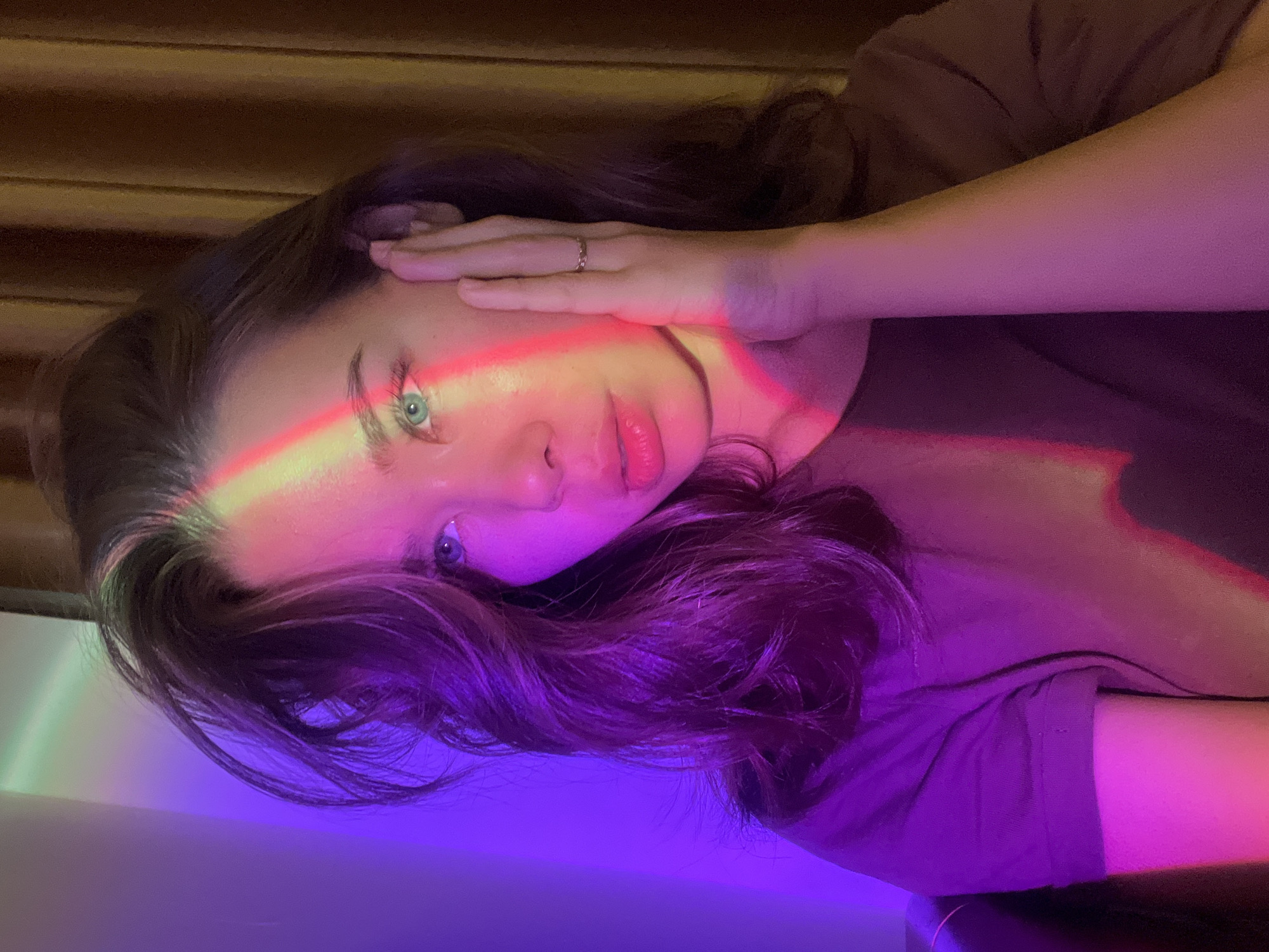 The image shows a young woman captured in a dramatic, artificially lit setting. She is oriented as if lying on her side, with her head resting, and her left hand positioned on her forehead. She has long, dark hair, and striking light-colored eyes, which appear blue or green. A prominent, vibrant band of colorful light, ranging from red and yellow to green and purple, cuts across her face, particularly illuminating her eye and lips. Her left hand, adorned with a simple ring on her ring finger, is also bathed in this spectrum of light.
She is wearing a dark-colored t-shirt. Her expression is calm and direct, looking towards the viewer. The background behind her head appears to be a textured surface, possibly wooden blinds or a similar wall treatment, with a darker hue. The lower part of the image shows more of the purple and green light cast onto a lighter, possibly white, surface. The distinct lighting suggests an artistic or experimental photography setup, possibly using a projection lamp to create the rainbow effect.
There is no discernible activity other than posing for the photograph. The time of day is not indicated, but the intense artificial lighting suggests an indoor setting during the evening or night. No specific location cues from "Nam Natural Heritage Area Trang, Vietnam" are visible within the image itself. There is no visible text.