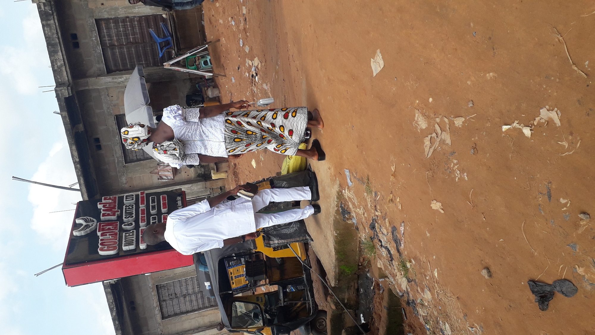 This media file depicts an outdoor scene in Nkpor, Nigeria. Two individuals are the primary subjects. A woman, dressed in a white traditional outfit with a vibrantly patterned headwrap, stands next to a man in a white traditional outfit. The man is holding a book or tablet. They appear to be standing on a dirt road next to a building with a red and black sign that reads "GOLDEN EAGLE HOTEL & SUITES LTD". The building looks to be under construction or in a state of disrepair, with exposed concrete and rudimentary window coverings. The ground is dirt and shows signs of litter. A yellow auto-rickshaw is parked nearby, partially visible. The sky is visible and appears cloudy. The lighting suggests it is daytime. The overall impression is that of a transactional or community interaction occurring on a busy street in a Nigerian town.