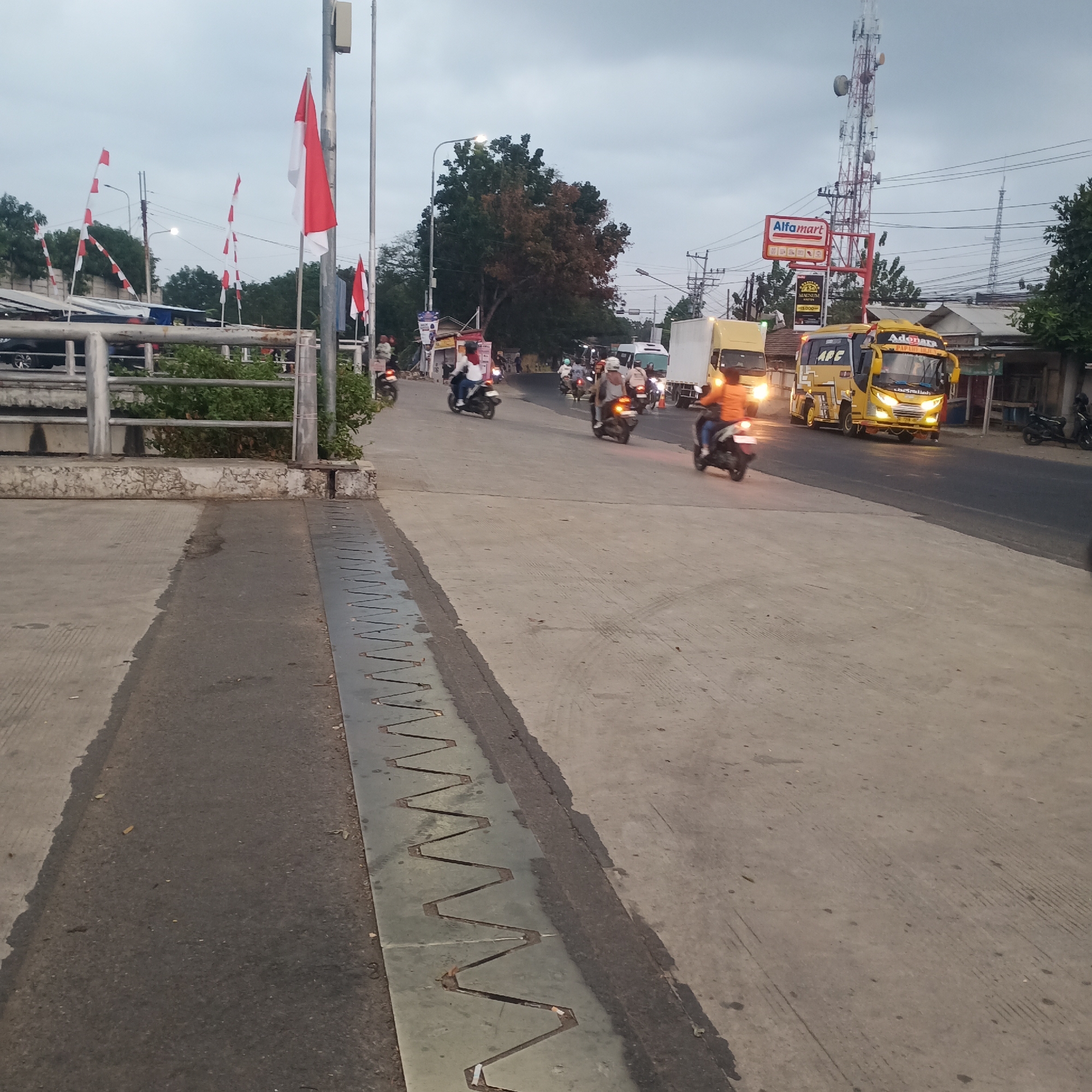 The image depicts a street scene in Balapulang, Indonesia, on an overcast day. A highway with multiple lanes of traffic is visible, populated by several vehicles including cars, trucks, and numerous motorcycles. Some motorcycles are being ridden by individuals wearing helmets, while others have passengers. The road is flanked by sidewalks, buildings, and vegetation. Indonesian flags are displayed prominently on poles along the side of the road, suggesting a national celebration or commemoration. A prominent red and white billboard with the "Alfamart" logo is visible, indicating a commercial area. The overall atmosphere is one of a busy, developing urban environment.