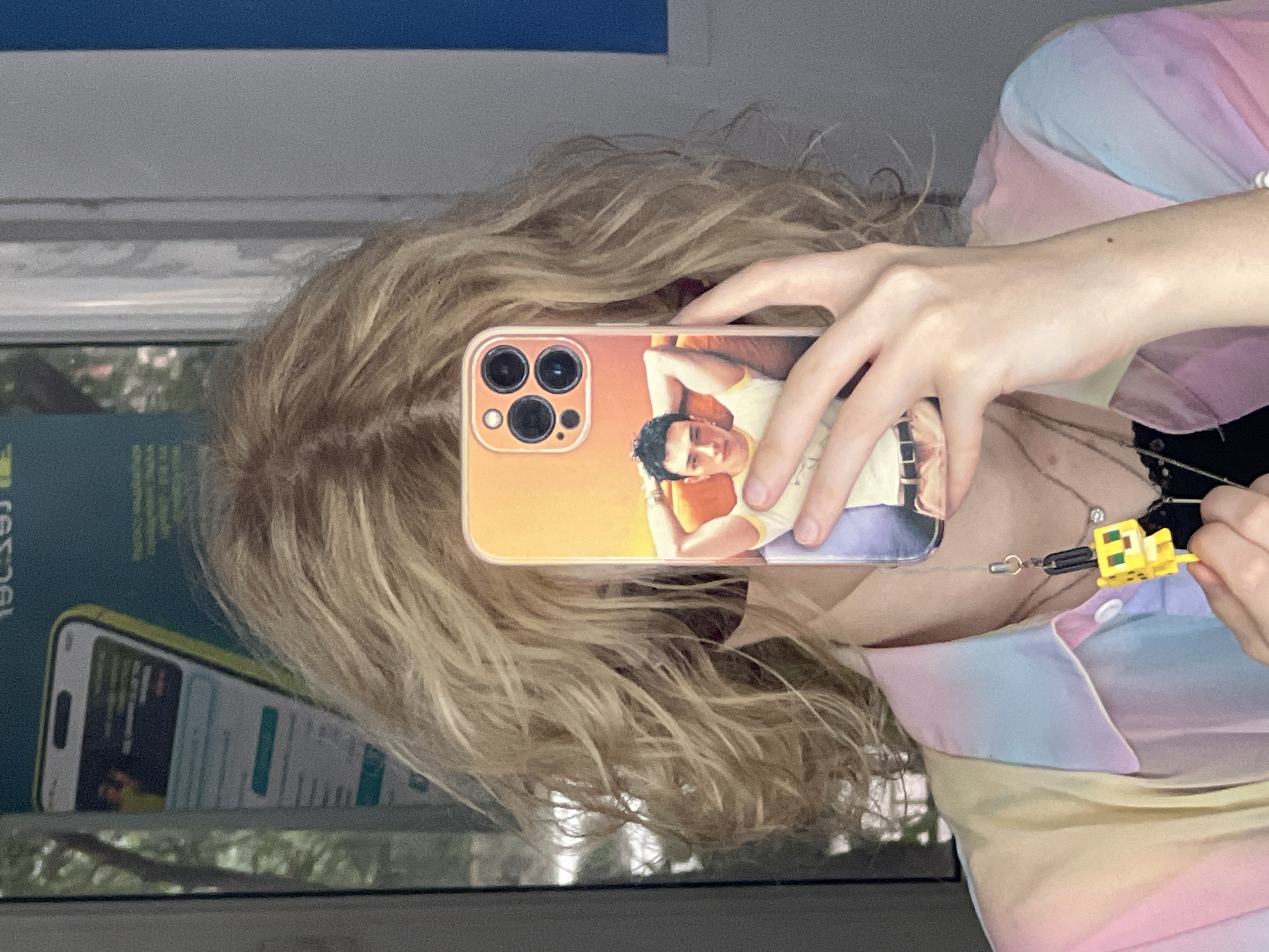 A person is taking a selfie in Almaty, Kazakhstan, holding up a phone with a picture of a man on the case. The person has blonde, curly hair and is wearing a multicolored pastel shirt. A yellow Minecraft-themed charm is attached to the phone. The background appears to be a window with reflections of trees and a street.