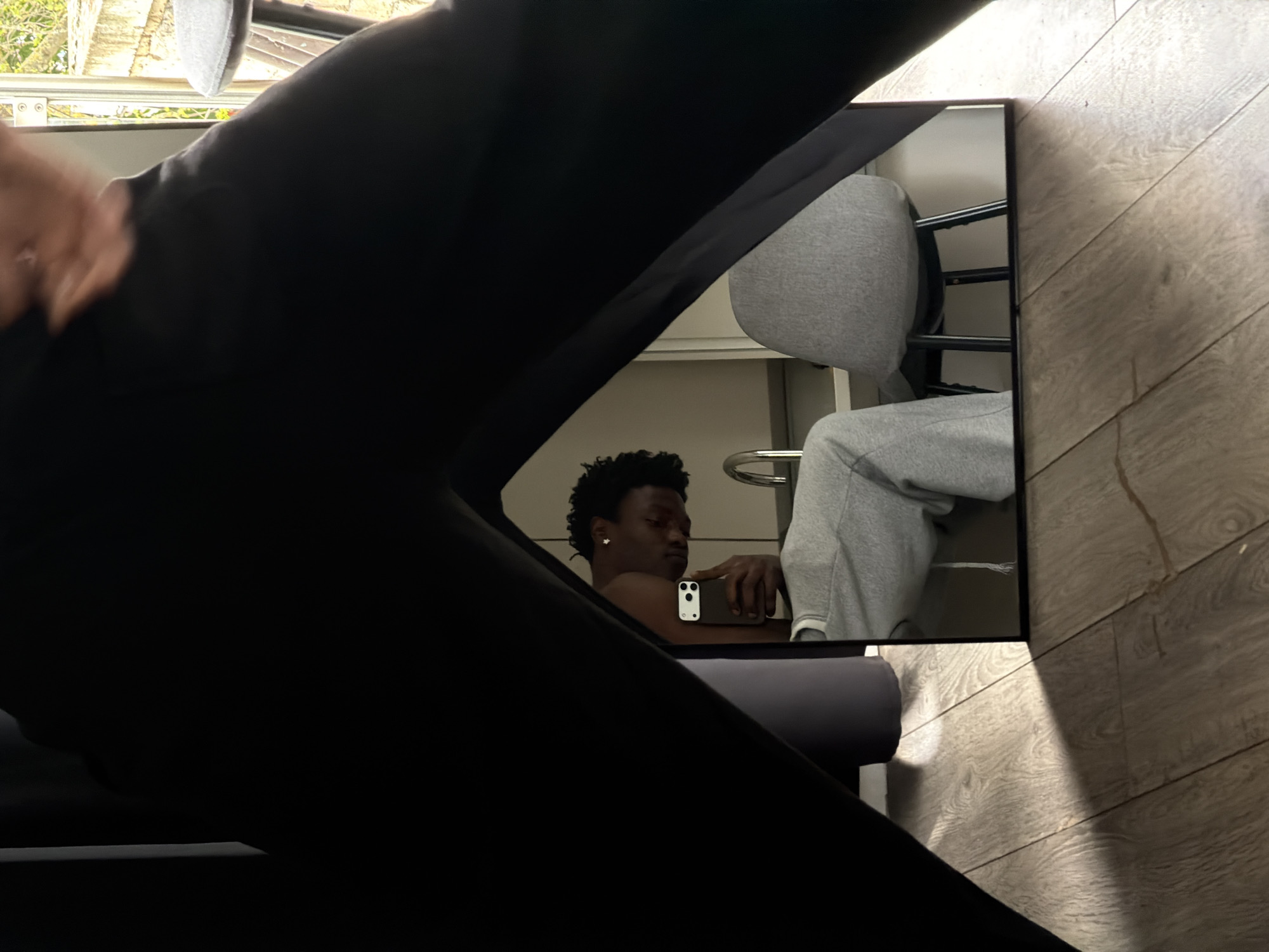 A low-angle, indoor photograph captures a reflection in a rectangular mirror, partially obscured by a large, dark, out-of-focus object that dominates the foreground, likely a leg clad in dark trousers or part of a piece of furniture.
In the mirror's reflection, the primary subject is a young Black man with short, dark, curly hair and a small star-shaped earring in his left ear. He appears to be shirtless or wearing a dark top and is holding a white smartphone with two visible cameras in his right hand, looking towards the device. To his right, also within the reflection, are the lower body and legs of another person, dressed in light grey sweatpants and a matching hoodie, seated on a chair with a black metal frame.
The surrounding room features light-colored wooden floorboards, with a prominent shadow cast across them to the right of the mirror, suggesting a direct light source. In the very top-left corner of the image, beyond the dark foreground and through a window, some greenery and what appears to be an old stone structure or building are visible.
The scene suggests a casual, relaxed home environment during the daytime, likely in London, United Kingdom, given the provided context. There is no visible text in the image.