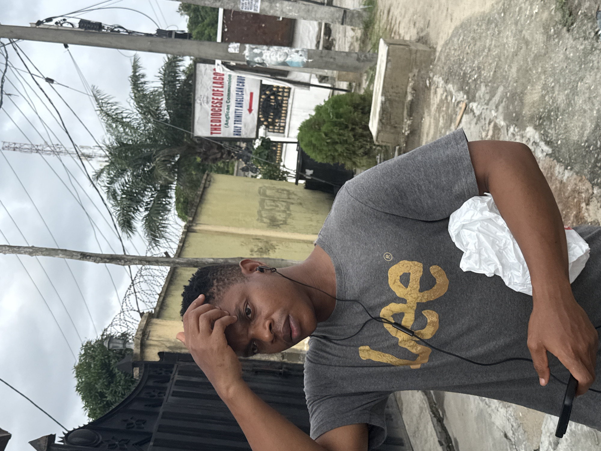 A medium shot captures a young man in Ado, Nigeria. He's wearing a gray T-shirt with a gold logo. He holds a smartphone in one hand and rests the other on his forehead. Earphones are visible. In the background is a sign for "THE DIOCESE OF LAGOS" and a direction to Holy Trinity Anglican Church. A palm tree is partially visible, along with utility poles, a walled building topped with barbed wire, and a metal gate. The sky is overcast.