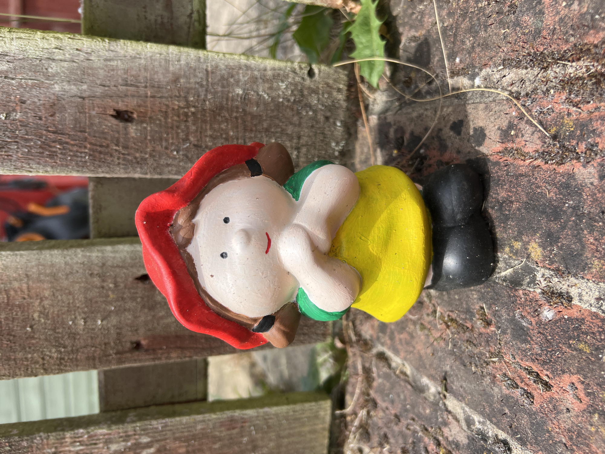 A ceramic gnome figurine is positioned on a weathered brick surface, with a wooden fence to the left. The gnome is painted with a red hat, white face, black eyes, red smile, brown hair, green collar, yellow body, and black boots. The background shows some greenery and glimpses of other structures. The location is Basingstoke and Deane, United Kingdom.