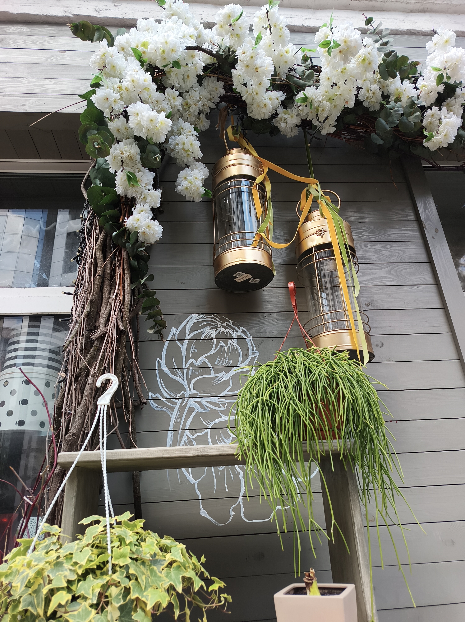 The image shows a decorated exterior wall, likely in Kyiv, Ukraine, though there are no specific Ukrainian identifiers other than the language spoken there. The wall is wood, painted grey, and decorated with white floral accents around what appears to be a window on the upper left. A branch or vine structure, adorned with white flowers and greenery, forms an arch across the top. Two gold-colored lanterns with clear glass sides are suspended from the floral arrangement, each decorated with ribbons in yellow and green tones. Below the arch, a simple wooden shelf supports two hanging plants. The left plant has variegated green and yellow leaves, hanging from a white plastic hook. To the right, a green plant with slender, stem-like foliage is also potted. A simplified white outline of a flower is painted on the wall between the vine structure and shelf. The bottom right corner shows a white square planter with a plant inside. The overall aesthetic is decorative and possibly intended for a commercial storefront.