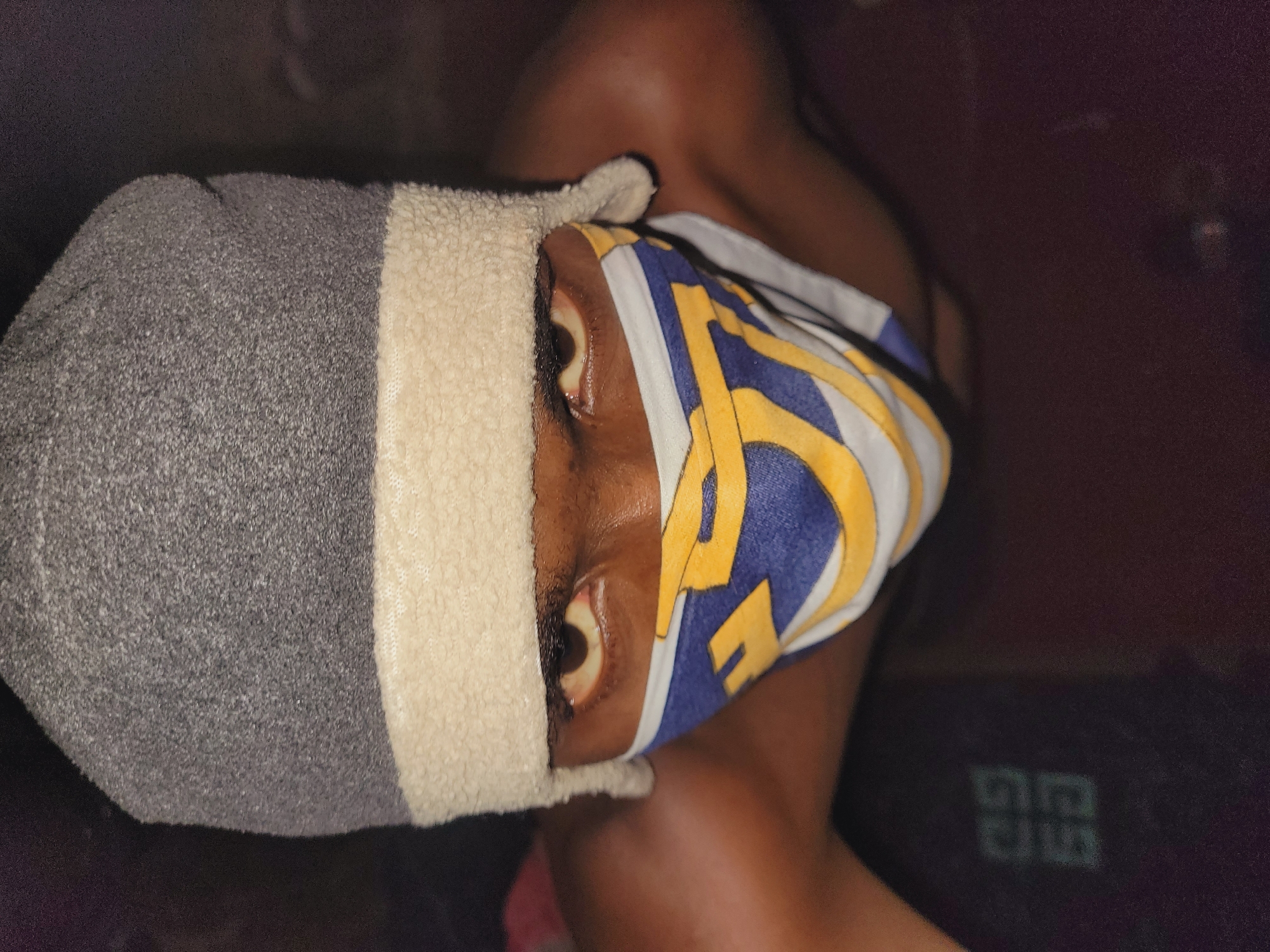 A close-up, high-angle shot shows a person wearing a grey beanie with a cream-colored trim and a patterned face mask with yellow and blue designs. The individual's eyes are visible and appear to be looking directly at the camera. The background is dark and out of focus, with a faint, blurry logo visible on a dark surface in the lower left quadrant of the image. The scene is not identifiable as being in Bukuru, Nigeria, or any other specific city.
