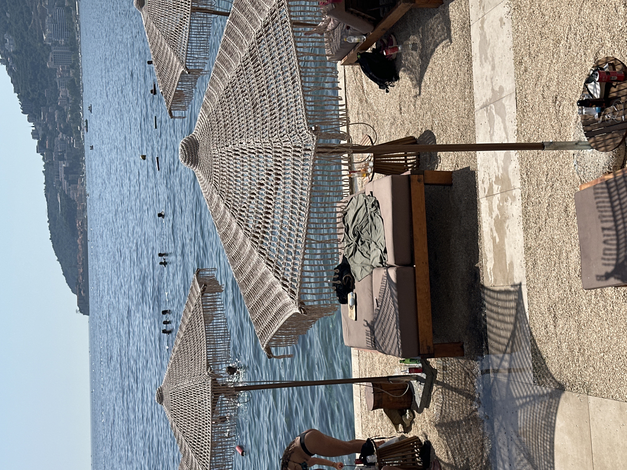 The image depicts a beach club in Budva, Montenegro, on a sunny day. The scene features the Adriatic Sea in the background with several people swimming. In the foreground, there are beach chairs and umbrellas, some with people relaxing on them. The ground is covered with gravel and stone tiles. There are several bottles and personal items scattered around the chairs. A woman in a black bikini is visible on the left side of the image, standing by the water. The overall atmosphere is one of leisure and relaxation at a seaside resort.