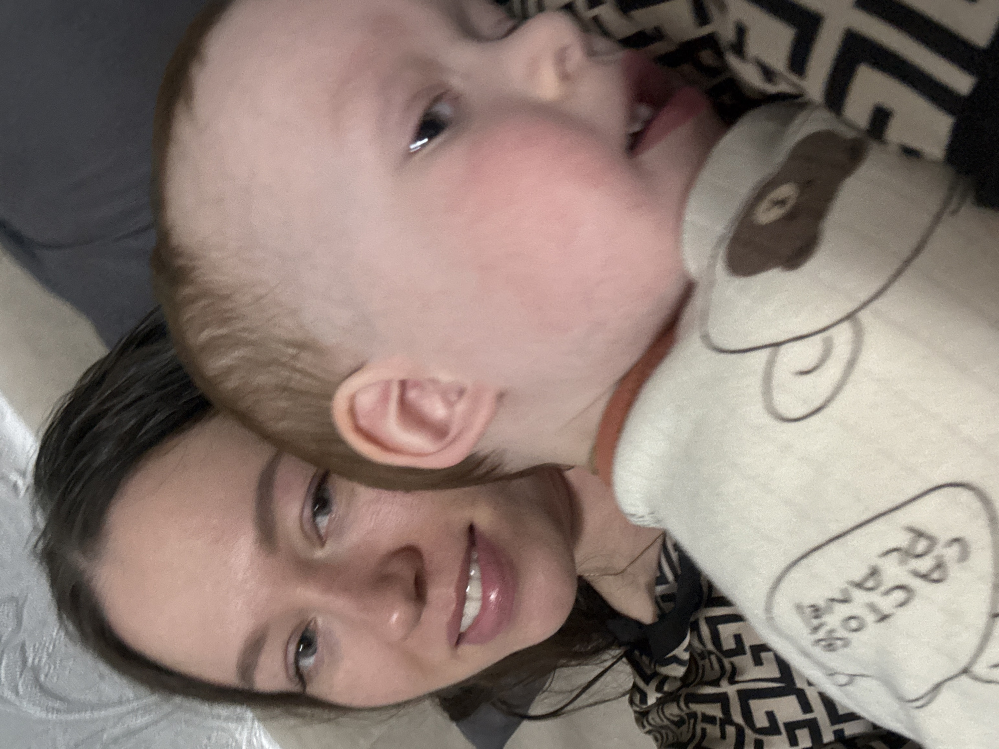 A woman with long brown hair and light skin is smiling and looking at the camera while holding a baby with fair skin. The baby is wearing a white outfit with a bear design on it. The woman is wearing a black and white patterned shirt. The image appears to be taken indoors. No details of the city of Ферма, Russia are present.