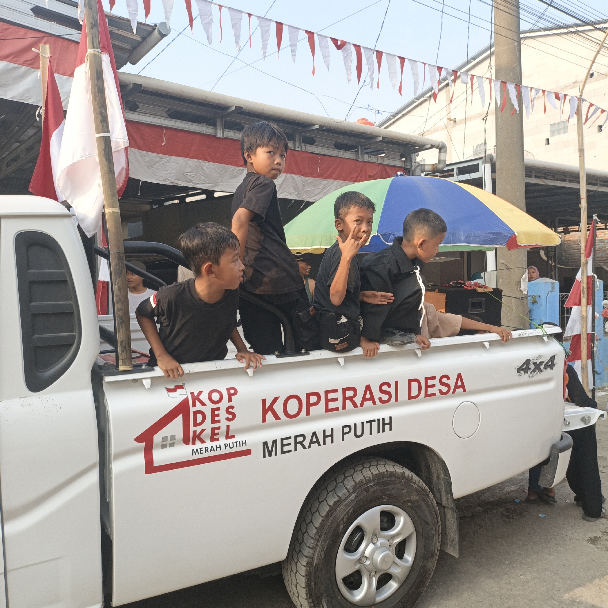 In Tanahbaya, Indonesia, a white pickup truck is decorated with red and white bunting and flags, likely for a celebration. Four young boys are visible in the truck bed. Three are wearing black t-shirts, and one is wearing a brown shirt. One boy in a black shirt is making a hand gesture. The truck has the words "KOP DESA KEL MERAH PUTIH" and "KOPERASI DESA MERAH PUTIH" printed on its side, along with a "4x4" emblem. A colorful umbrella is positioned behind the boys. In the background, buildings, power lines, and a few adults are present. The ground appears to be asphalt.