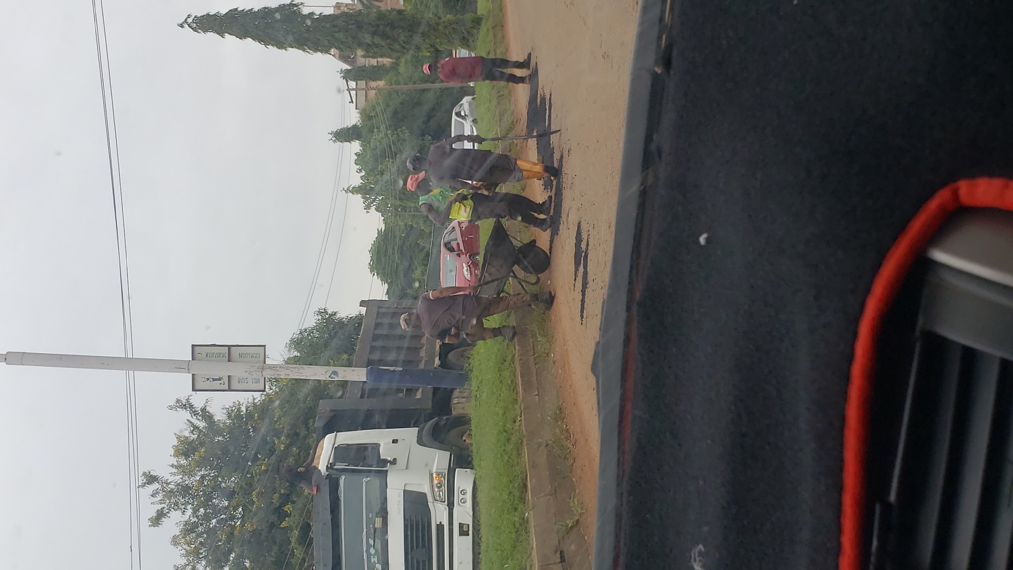 The image depicts a road construction scene in Unguwar Shanu, Nigeria, on an overcast day. In the foreground, the interior of a vehicle is visible, with a dark dashboard and red trim. The main focus is on the road and its surroundings. A white dump truck is parked on the side of the road, with a dark bed. Several construction workers are engaged in roadwork. One worker, wearing a camouflage uniform, is pushing a wheelbarrow filled with what appears to be asphalt. Another worker, in a yellow vest and red cap, is also near the wheelbarrow, assisting or supervising. A third worker, in a red shirt and cap, stands further down the road, holding a shovel. There are piles of asphalt on the road surface, indicating active repair or construction. A red car is parked behind the dump truck. The background features a grassy area, trees, and a tall, thin tree on the right side of the image. A tall, white utility pole with signs attached stands near the construction site. The sky is uniformly gray, suggesting cloudy weather.