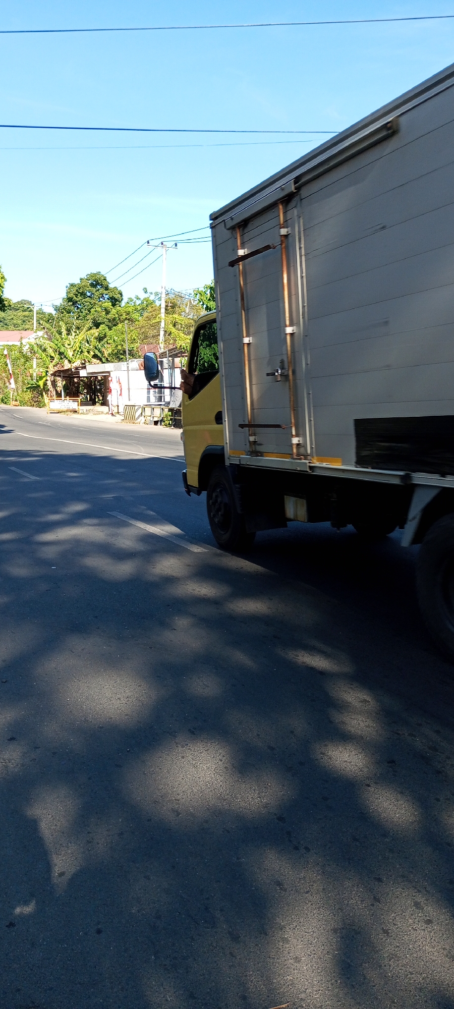 In Bulupabbulu, Indonesia, a yellow truck with a grey box trailer is driving on a paved road. The truck is positioned on the right side of the frame, with its trailer extending across the image. The road has white lane markings. In the background, there are trees, buildings, and utility poles with wires. The sky is clear and blue. Shadows of trees are cast on the road in the foreground.